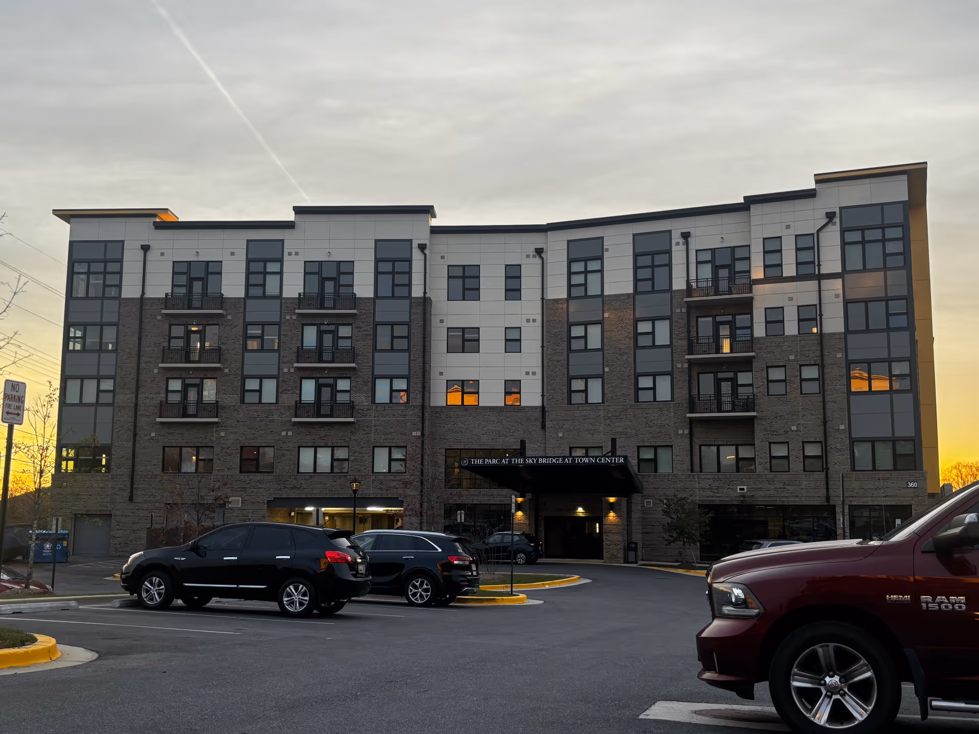 Front exterior of a multi-story senior living building with balconies and parked cars in the lot at sunset.