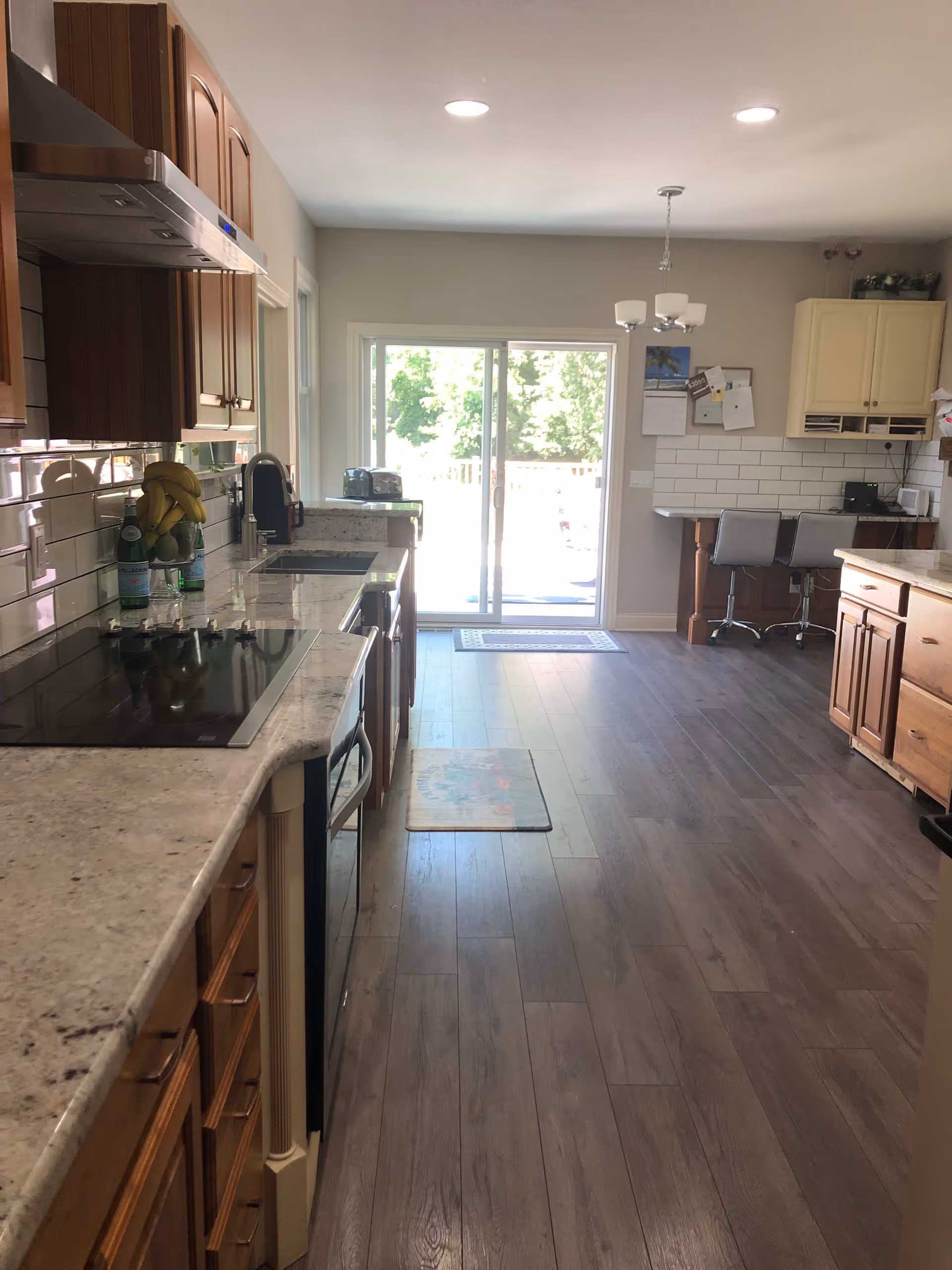 A bright kitchen with wooden cabinets and light-colored granite countertops. The kitchen features a stovetop, oven, sink, and various small appliances. There is a sliding glass door at the far end letting in natural light, and a small dining area with two chairs and a table against the wall. The floor is covered with wood-like laminate.