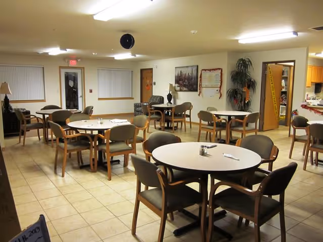 Interior view of a dining room in a senior living facility with multiple round tables and chairs arranged neatly. The room has tiled flooring, fluorescent ceiling lights, and windows with blinds. There is a door at the back, some wall decorations, and a small kitchen or serving area visible through an open doorway.