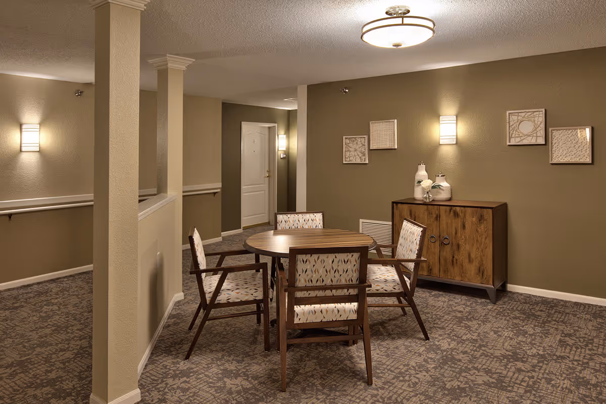 Carpeted communal seating area with a round wooden table, four upholstered chairs, a wooden sideboard, wall art, and soft wall lighting in a hallway.