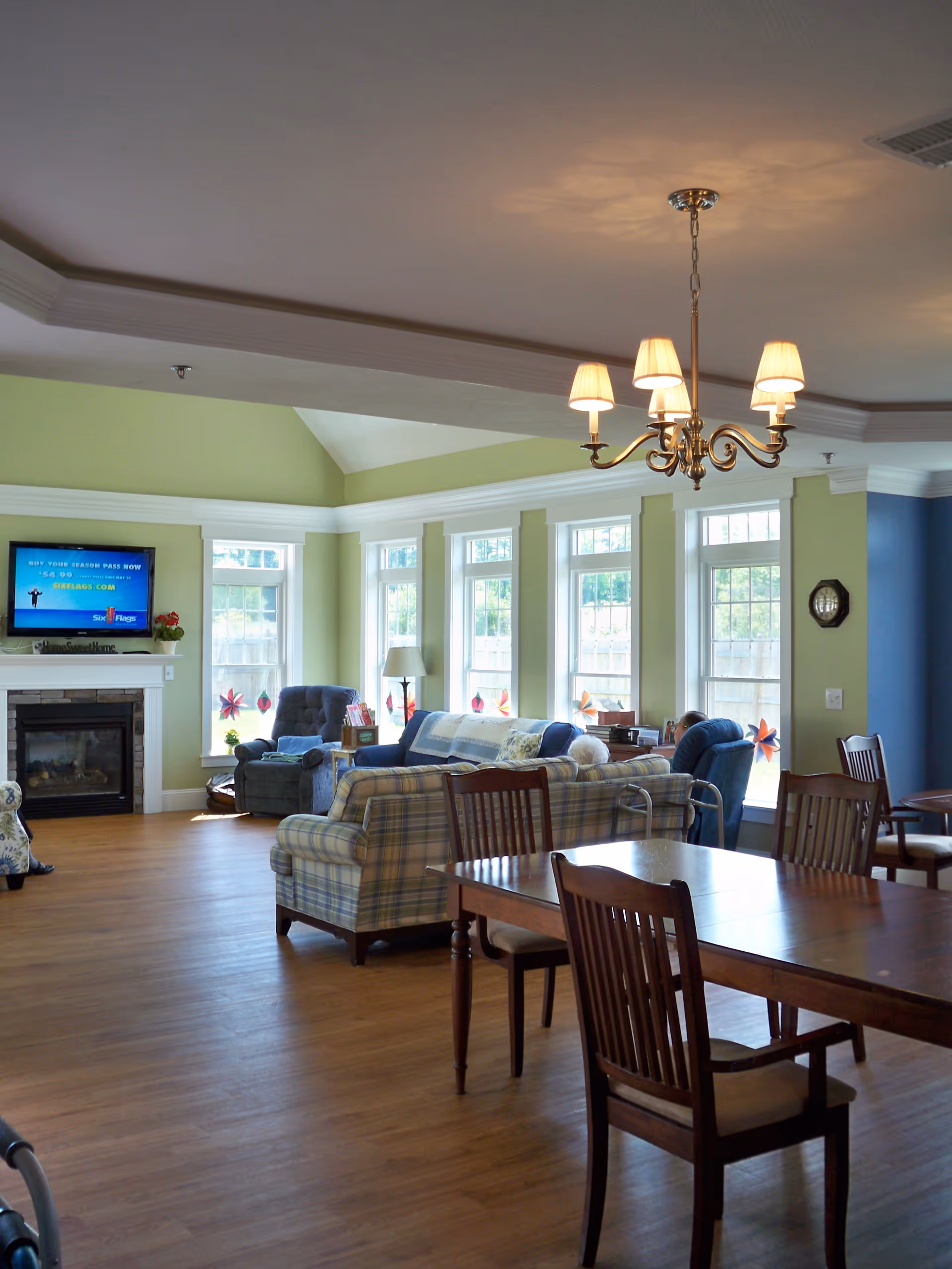 A bright and spacious living room area in a senior living facility with large windows letting in natural light. The room features multiple cushioned armchairs and sofas arranged around a fireplace with a flat-screen TV mounted above it. There is a wooden dining table with chairs in the foreground and a chandelier hanging from the ceiling. The walls are painted in light green and blue tones.