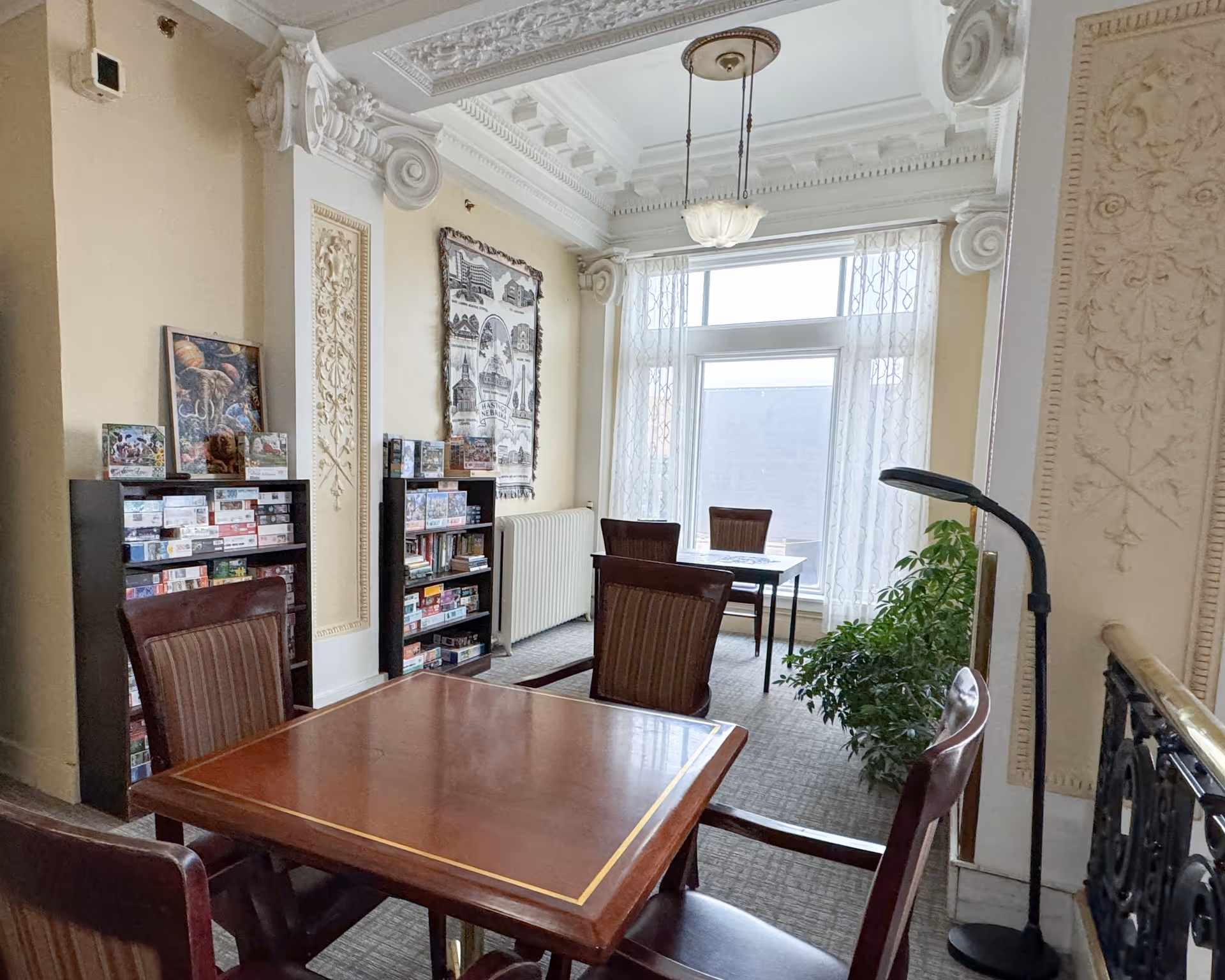 A cozy room with ornate ceiling moldings and decorative wall panels, featuring two wooden tables each surrounded by four wooden chairs with cushioned seats. Two black bookshelves filled with board games and books are against the wall, and a large window with sheer curtains lets in natural light. A green potted plant and a black floor lamp are also visible.