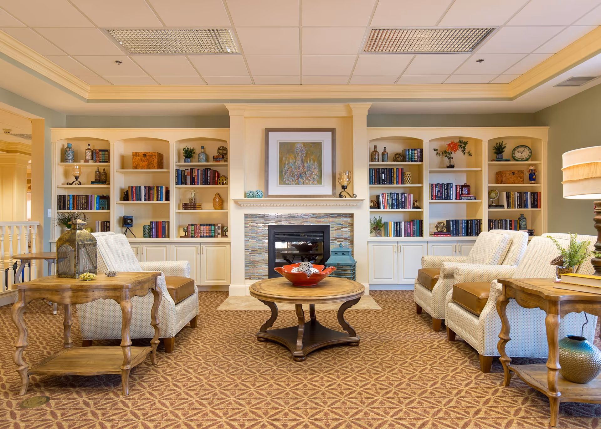 A cozy living room area with two armchairs and a wooden coffee table in the center. Behind the seating area is a fireplace with a framed painting above it, flanked by built-in bookshelves filled with books and decorative items. The room has a patterned carpet and soft lighting.
