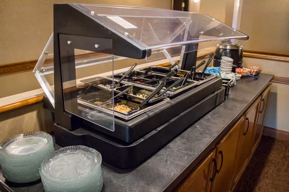 Buffet-style food service station with a clear sneeze guard, multiple food pans and serving utensils on a countertop with stacks of plates.