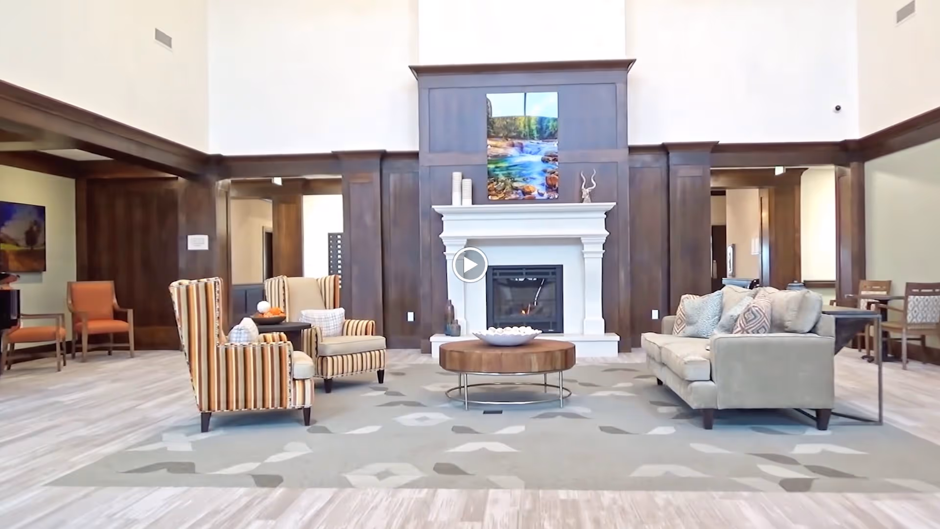 A spacious and well-lit living room area with a large fireplace centered on a dark wood-paneled wall. In front of the fireplace, there is a round wooden coffee table on a patterned rug. Two striped armchairs with pillows are positioned on the left side of the table, and a beige sofa with multiple cushions is on the right. Additional seating and tables are visible in the background.