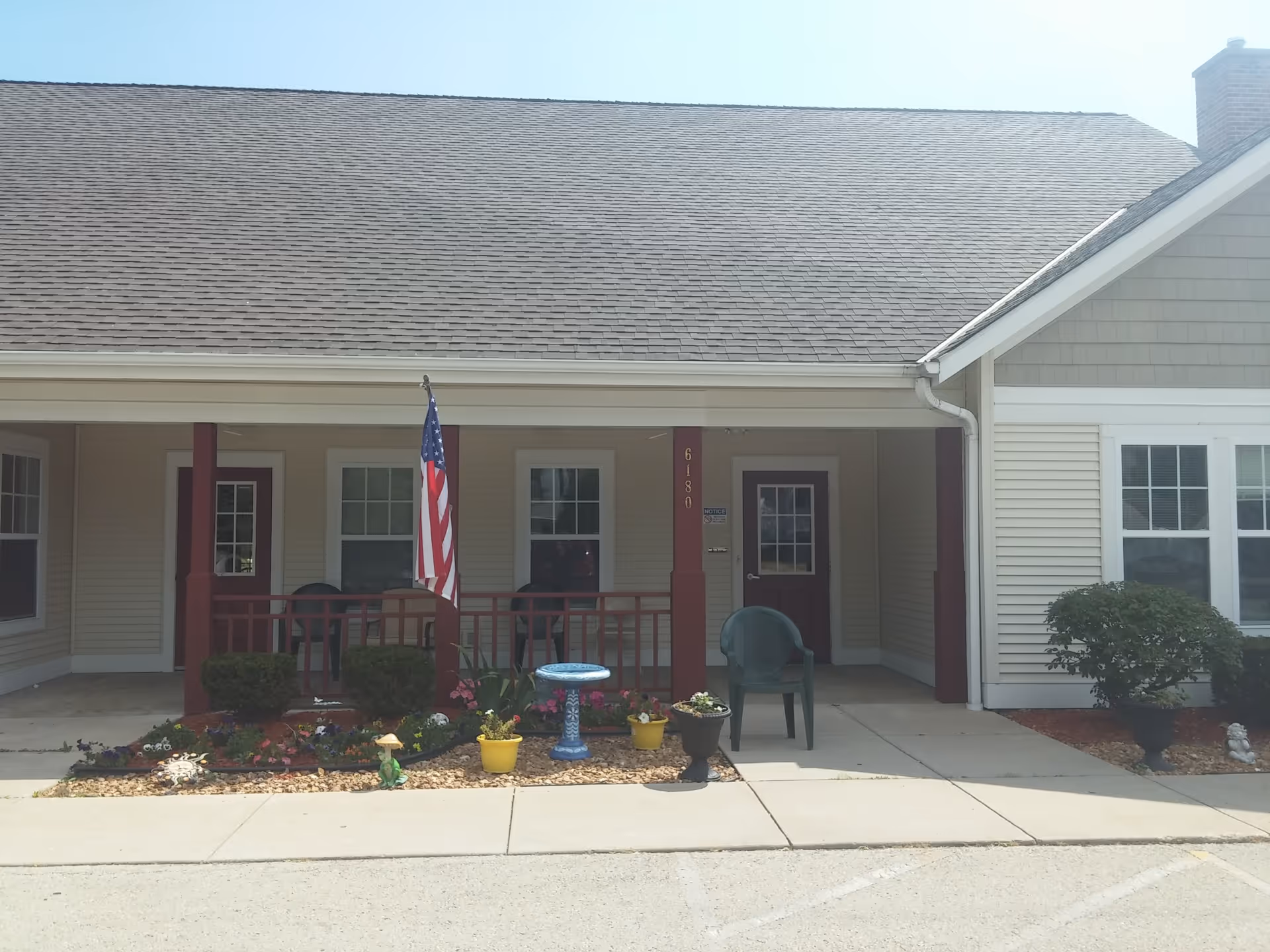 Front exterior view of a single-story building with beige siding and a gray shingled roof. There is a covered porch with red pillars and railing, an American flag mounted on one pillar, two green chairs, and a small garden with colorful flowers and decorative pots in front. The building has two doors and several windows.