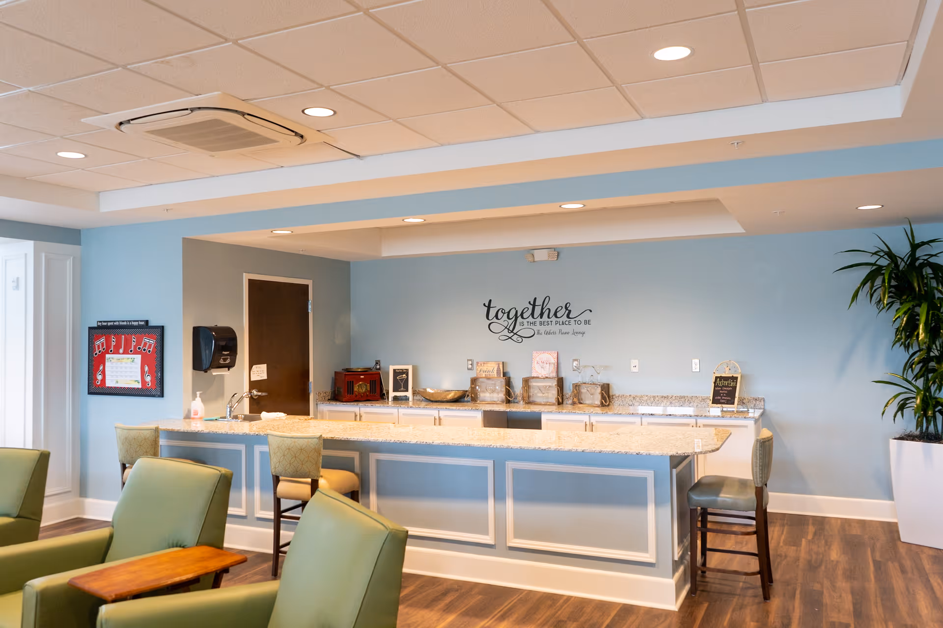 A bright and welcoming common area with a long counter and bar stools, light blue walls, and comfortable green armchairs. The wall behind the counter has a decorative quote that reads 'together is the best place to be.' There is a potted plant on the right side and a framed musical-themed picture on the left wall.