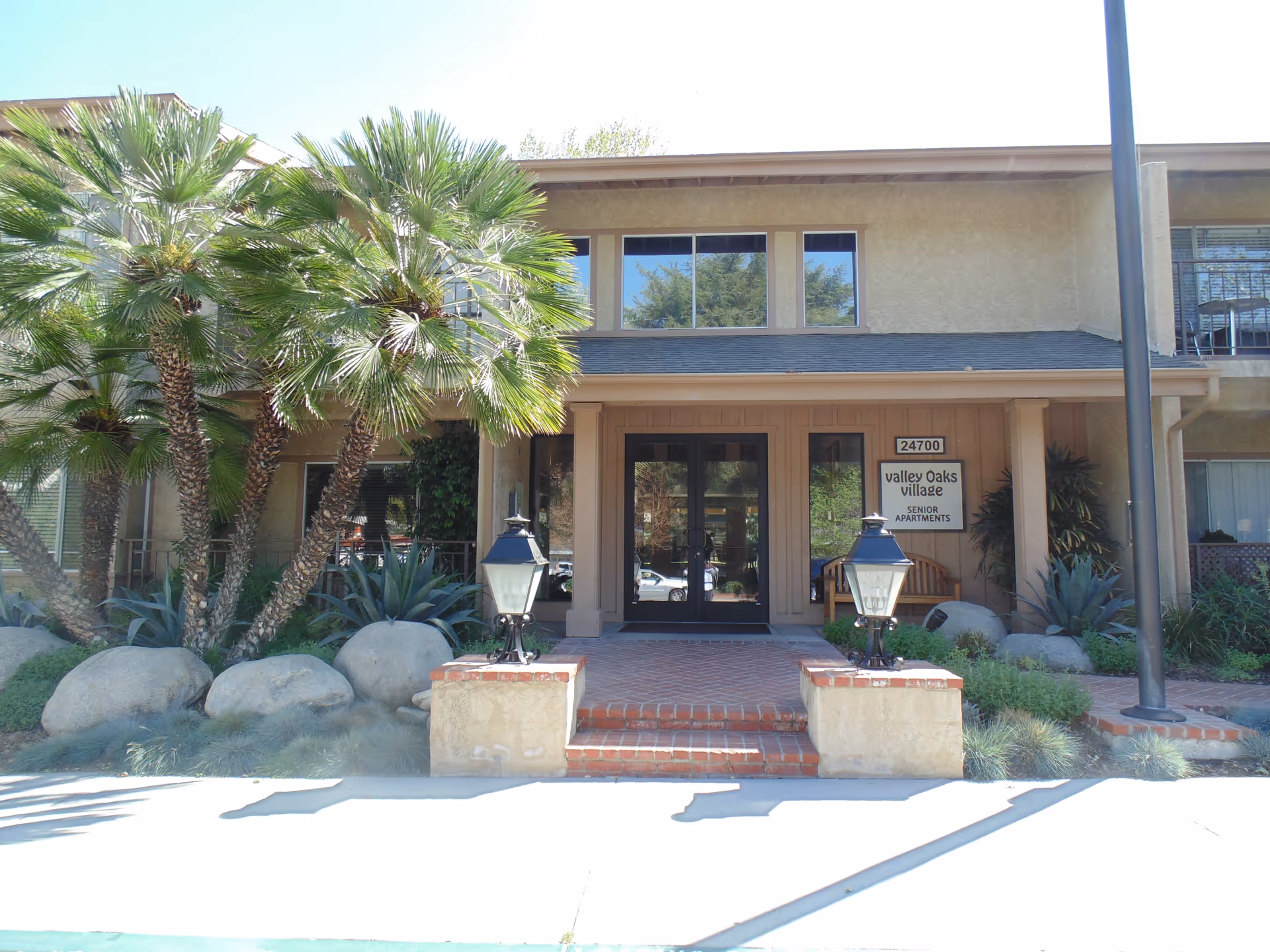 Front entrance of Valley Oaks Village senior apartments building with double glass doors, brick steps, two lantern-style lights on either side of the steps, palm trees, large rocks, and various plants in the landscaping.