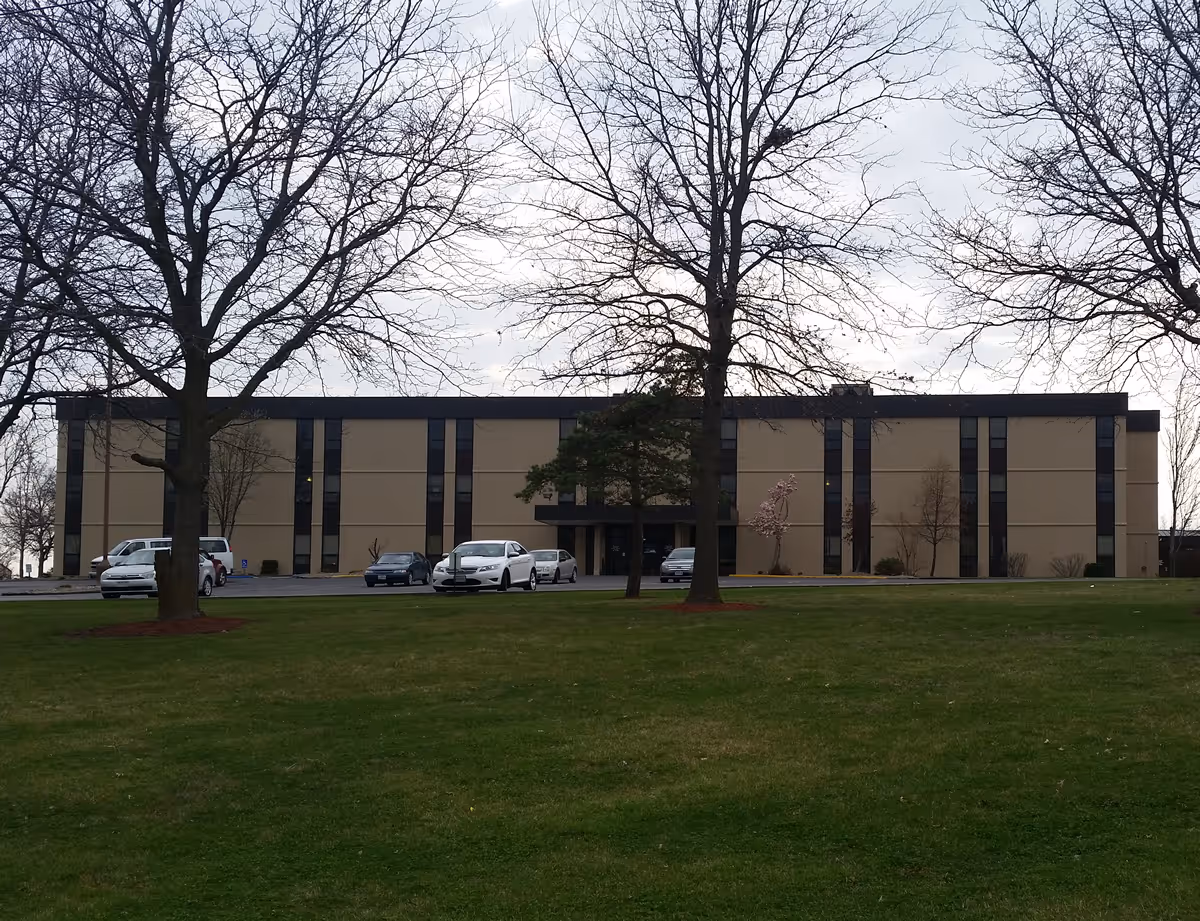 Front exterior of a three-story beige institutional building with vertical dark windows, parked cars, leafless trees, and a grassy lawn.