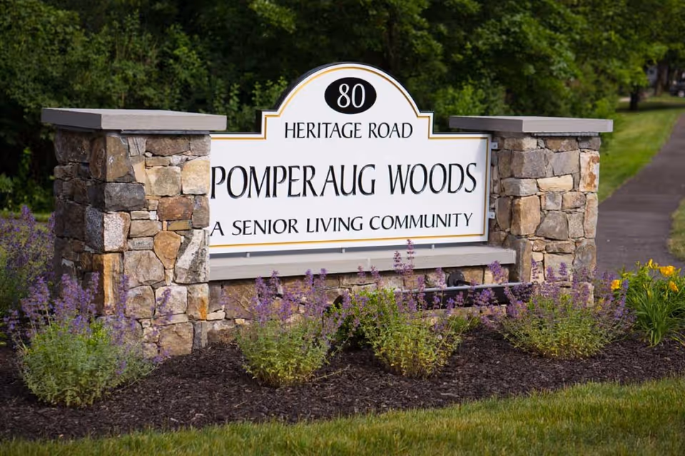 Stone signpost with the address '80 Heritage Road' and the name 'Pomperaug Woods, A Senior Living Community' surrounded by purple and yellow flowers and greenery.