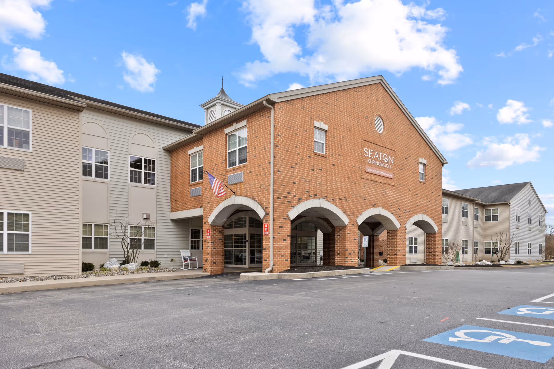 Exterior view of Seaton Springwood facility showing a two-story building with a brick entrance featuring three arches, an American flag, and multiple windows. The parking lot in front includes designated handicap parking spaces under a partly cloudy sky.