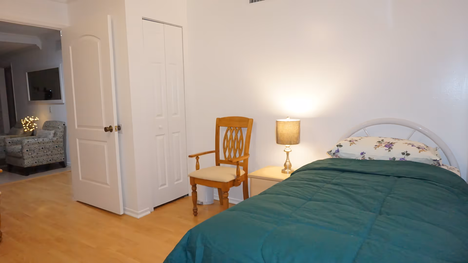 A bedroom with a single bed covered with a green comforter and a floral pillow. Next to the bed is a wooden chair and a small nightstand with a lit table lamp. The room has light-colored wooden flooring and white walls. An open door leads to another room with an armchair and a TV mounted on the wall.