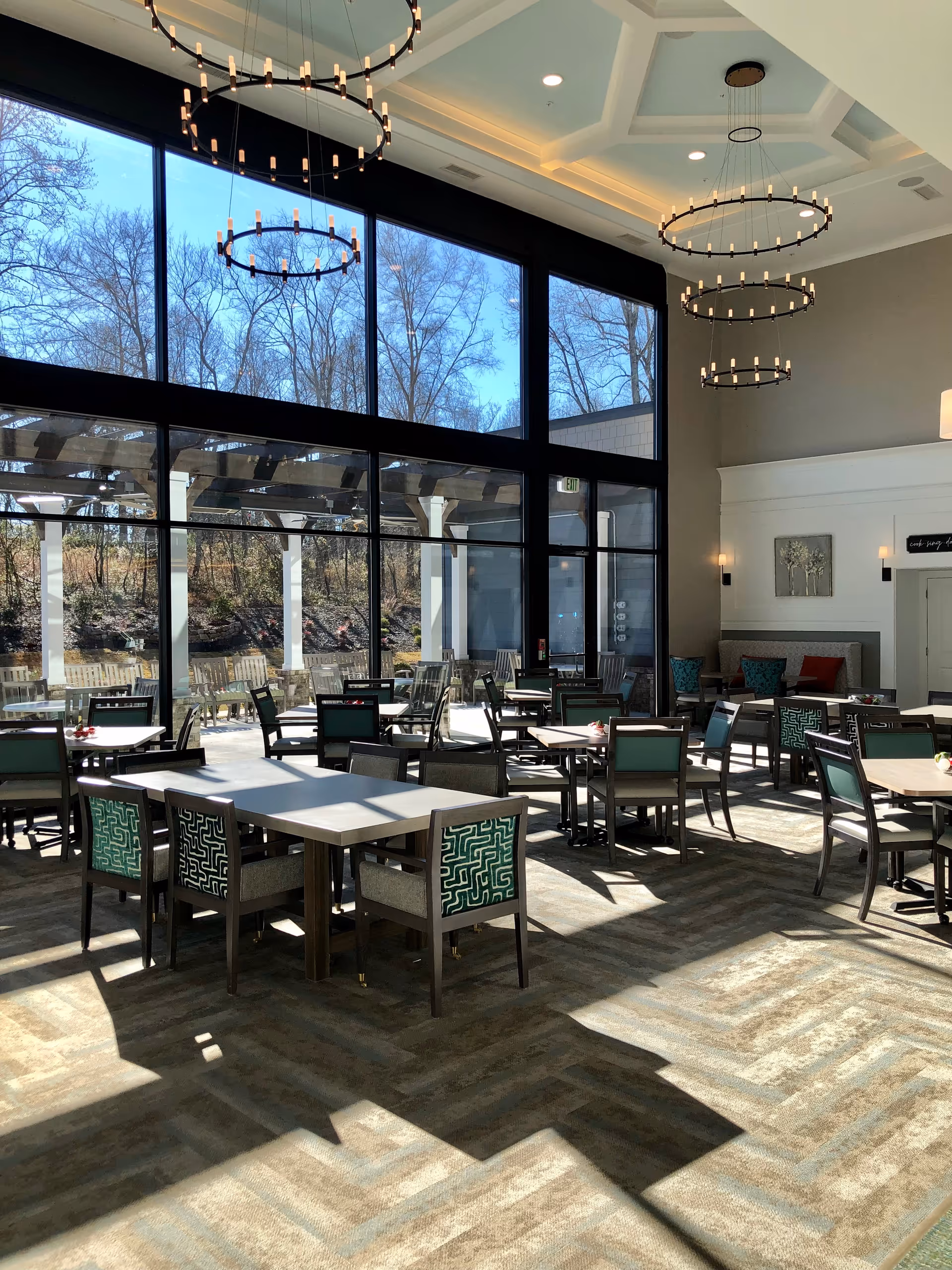 A bright dining room with large floor-to-ceiling windows letting in natural light. The room features multiple tables with chairs that have green patterned upholstery. Outside the windows, there is a patio area with wooden benches and a pergola. Modern circular chandeliers hang from the ceiling, and there are wall sconces and artwork on the walls.