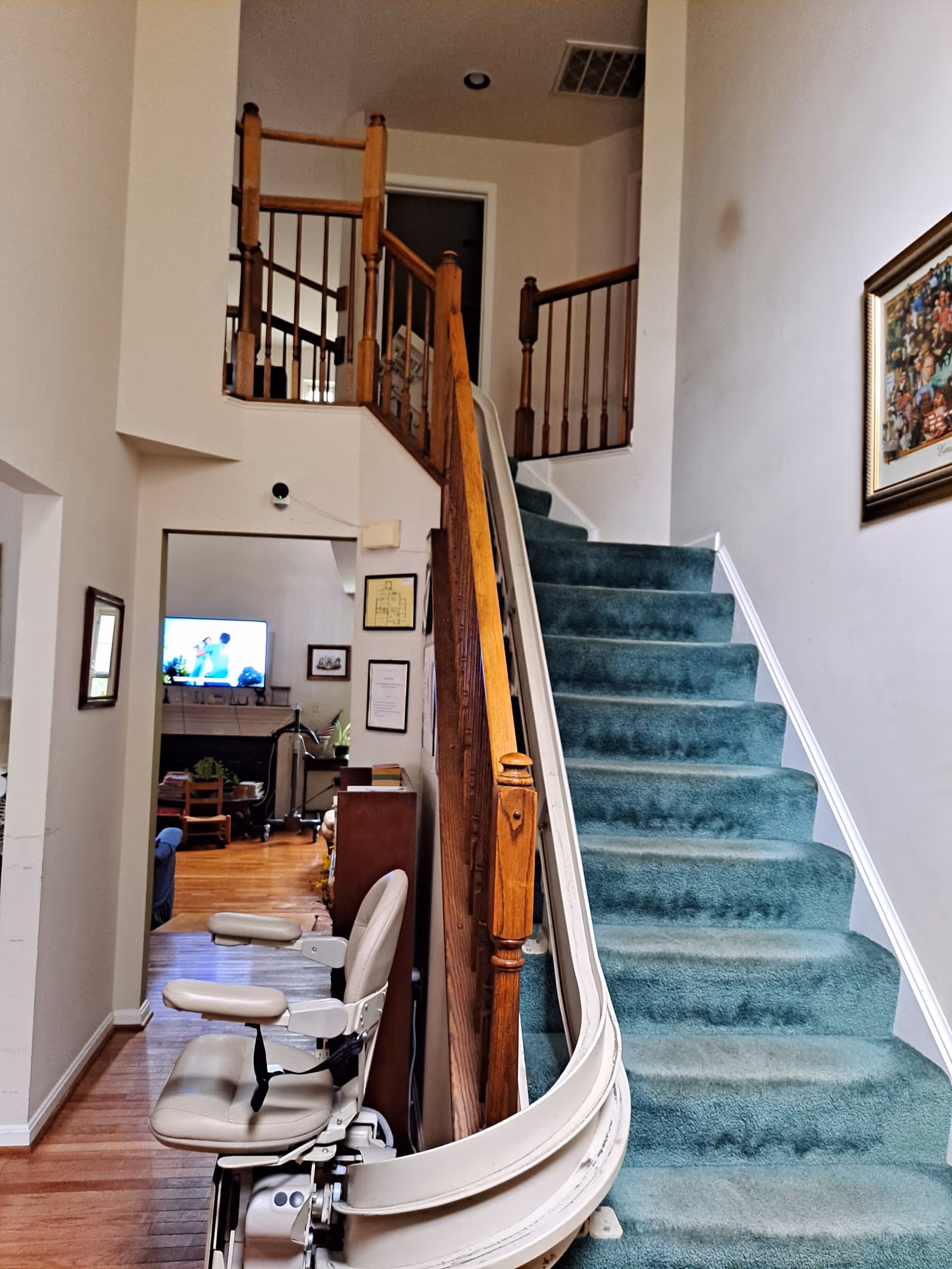Interior view of a home with a staircase covered in green carpet. A stairlift is installed on the right side of the stairs. The staircase has wooden railings, and there is a hallway leading to a living room area with a television and furniture visible in the background. A framed picture hangs on the wall to the right of the stairs.