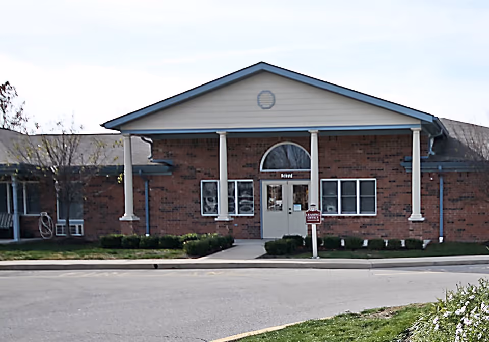 Front exterior view of a single-story brick building with white columns supporting a triangular pediment above the entrance. There are windows on either side of the double door entrance, and a small sign is visible near the door. The surrounding area includes a paved driveway and some greenery.