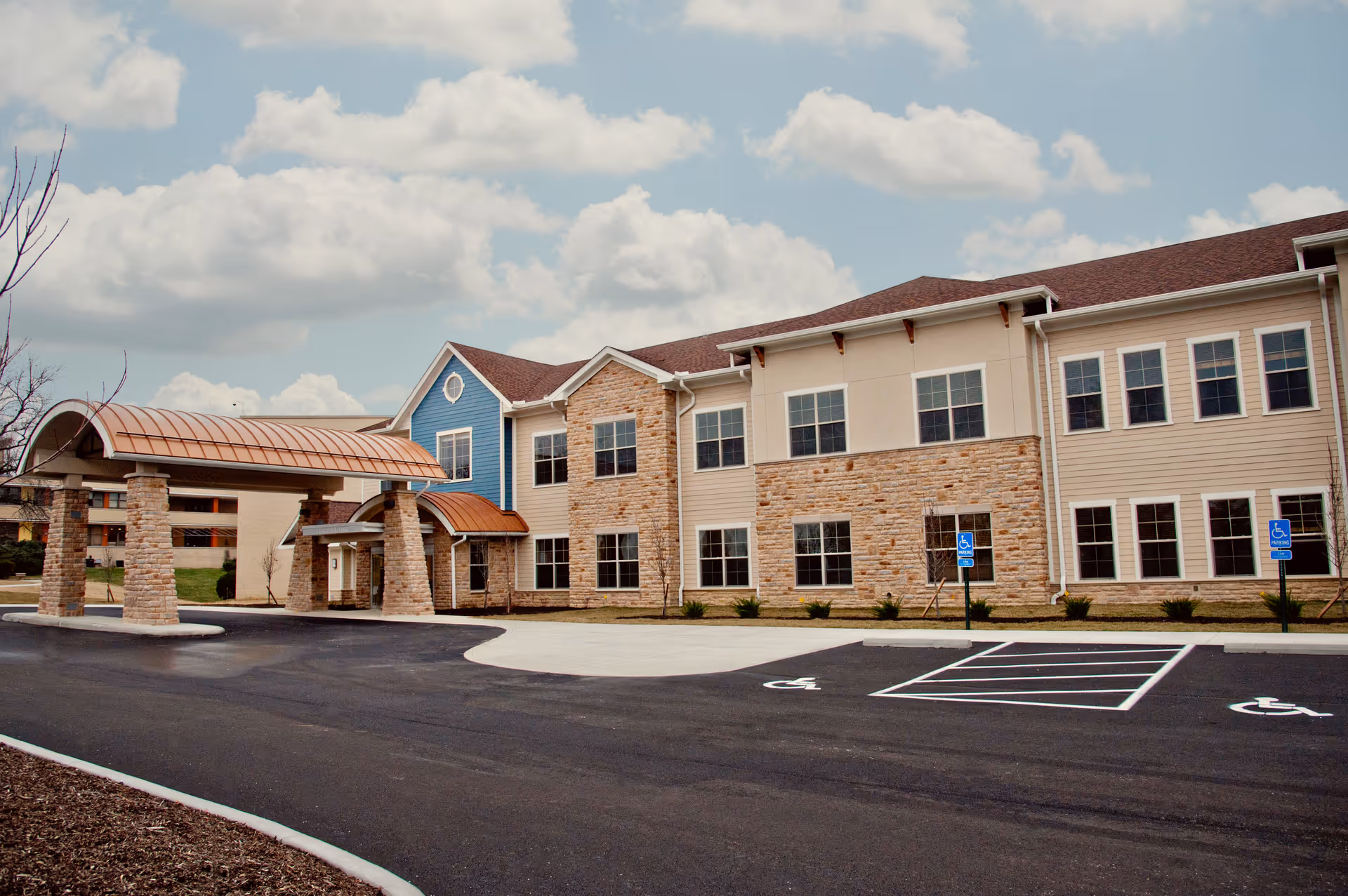 Exterior view of a two-story senior living facility building with a covered entrance supported by stone pillars. The building features a mix of stone and siding on the facade, multiple windows, and a parking area with handicap spaces in front. The sky is partly cloudy.