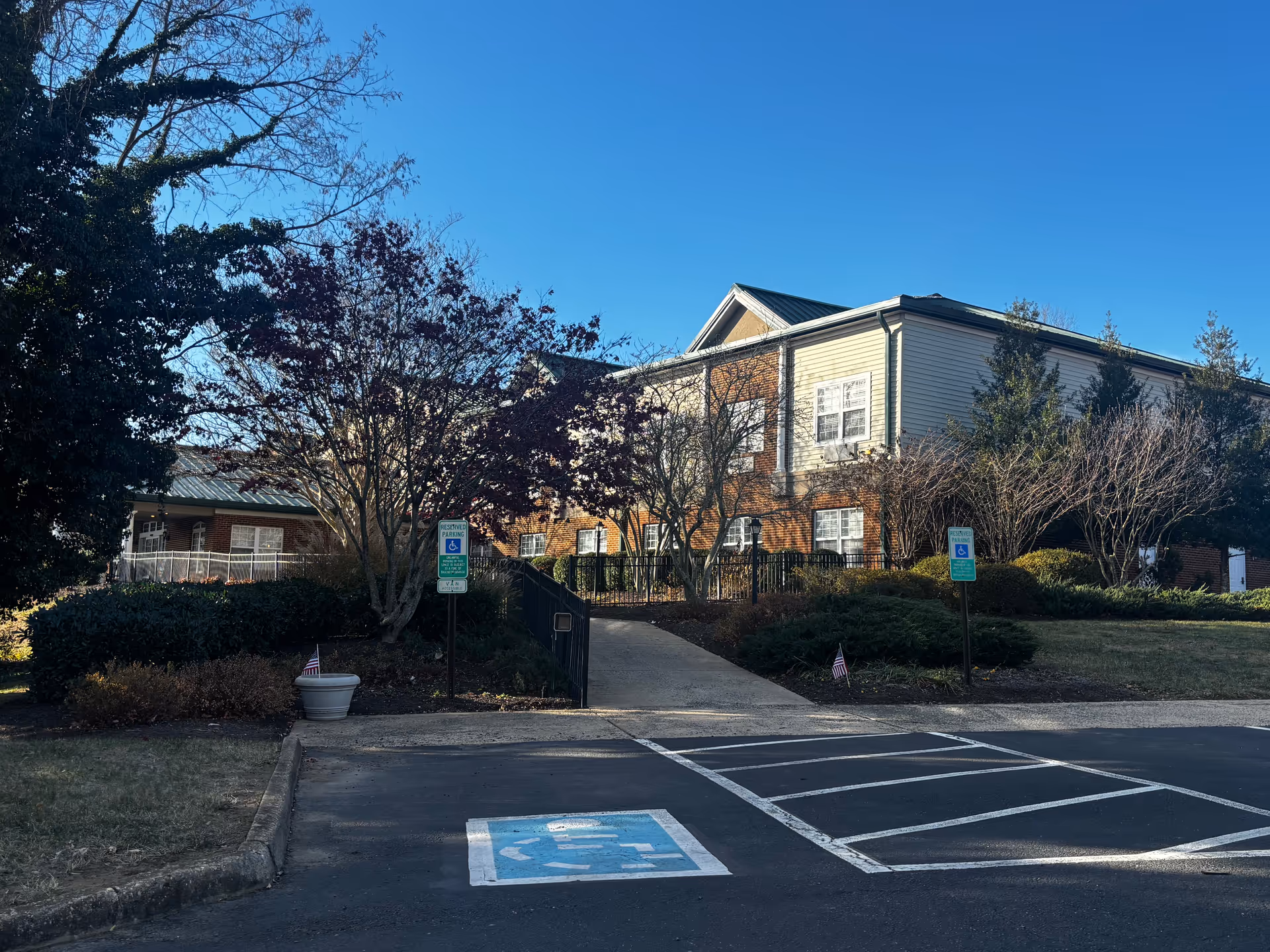 Exterior view of a senior living facility building with a brick and beige siding facade, surrounded by trees and bushes. There is a paved parking area in the foreground with two reserved handicapped parking signs and a wheelchair accessible parking space marked on the ground. The sky is clear and blue.