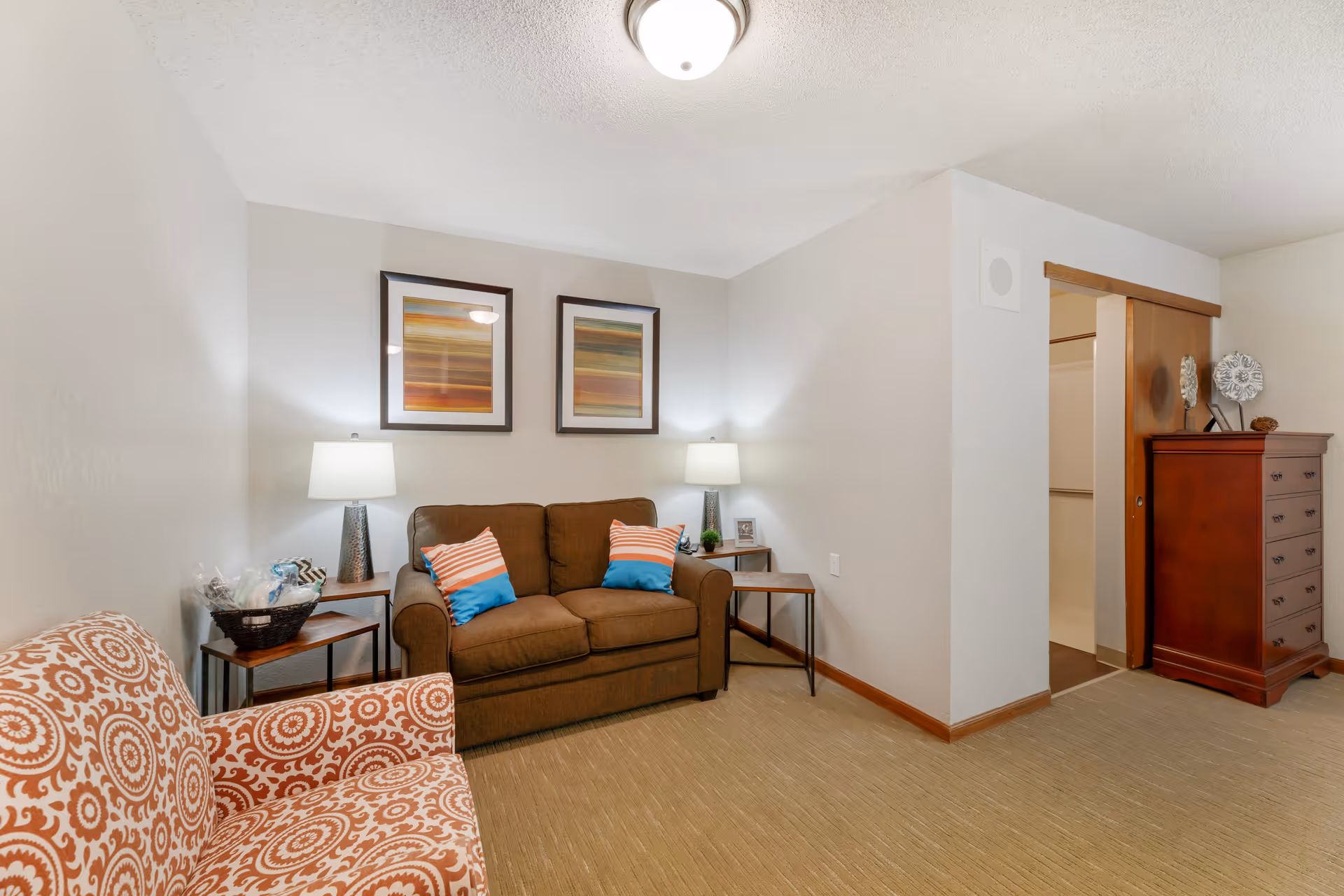A cozy living room with a brown loveseat adorned with four colorful pillows, two side tables each with a lamp, an orange patterned armchair, two framed abstract artworks on the wall, and a wooden dresser near a sliding door leading to a closet.