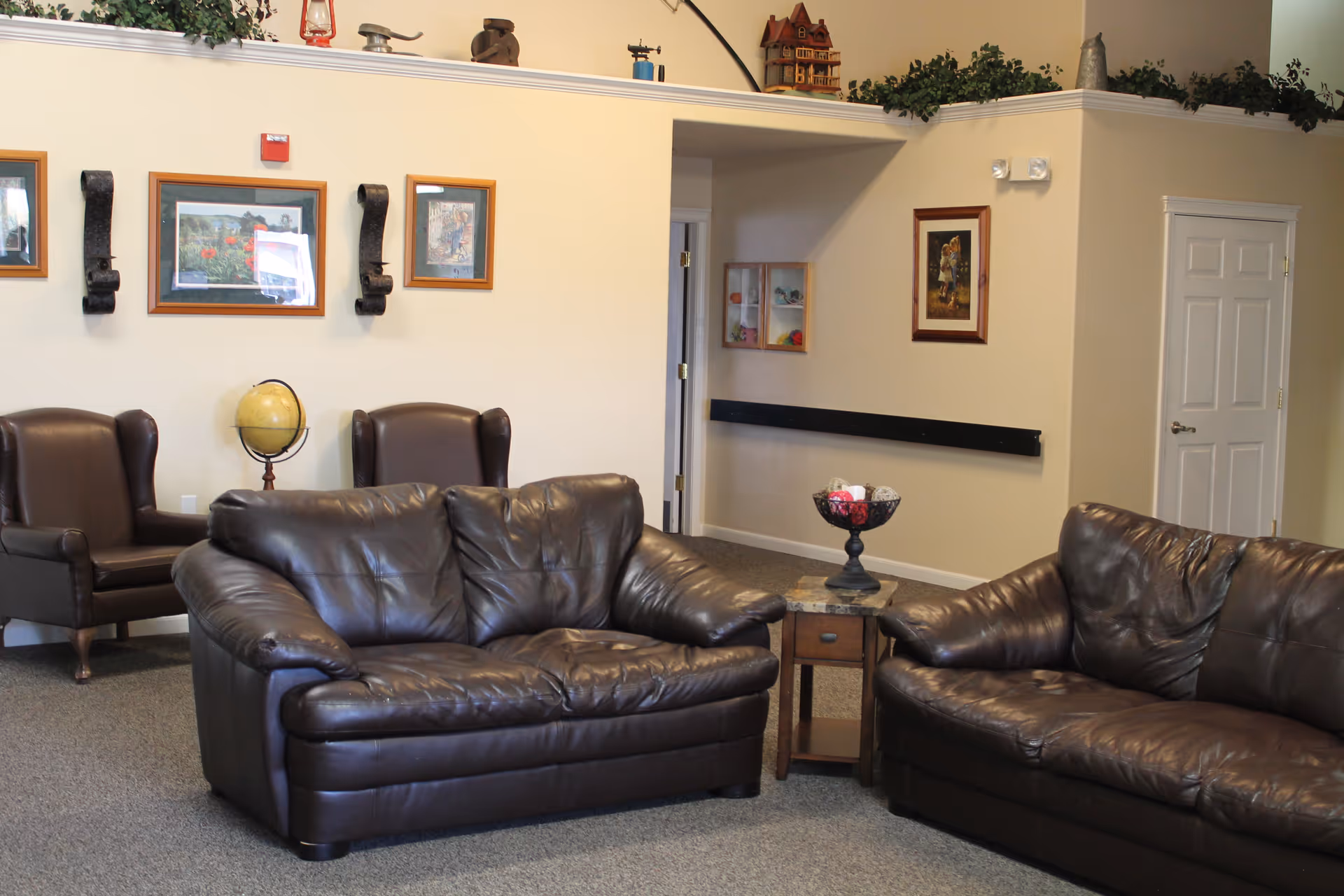 A communal living room with brown leather sofas and armchairs, framed artwork on the walls, and decorative plants above the ledge.