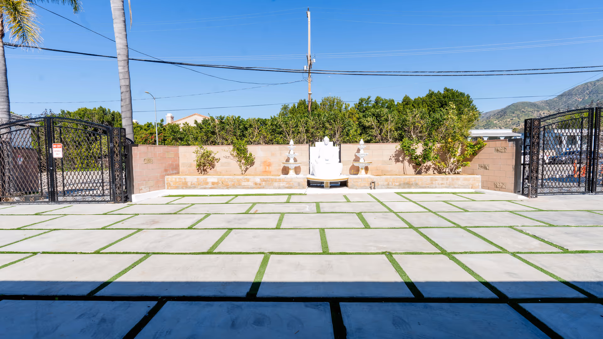 Outdoor courtyard area with large concrete tiles separated by green grass strips, a brick wall with small plants, two black metal gates on either side, and a white Buddha statue centered against the wall under a clear blue sky.
