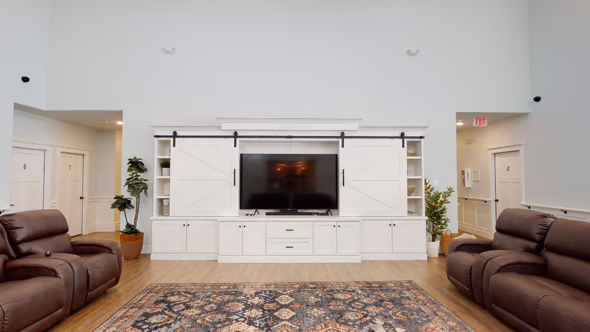 A bright living room area with two brown leather recliners facing a large flat-screen TV mounted on a white built-in cabinet with sliding barn doors. The room has light-colored walls, wood flooring, a patterned area rug, and potted plants on either side of the TV unit. Doors labeled with numbers are visible in the background.