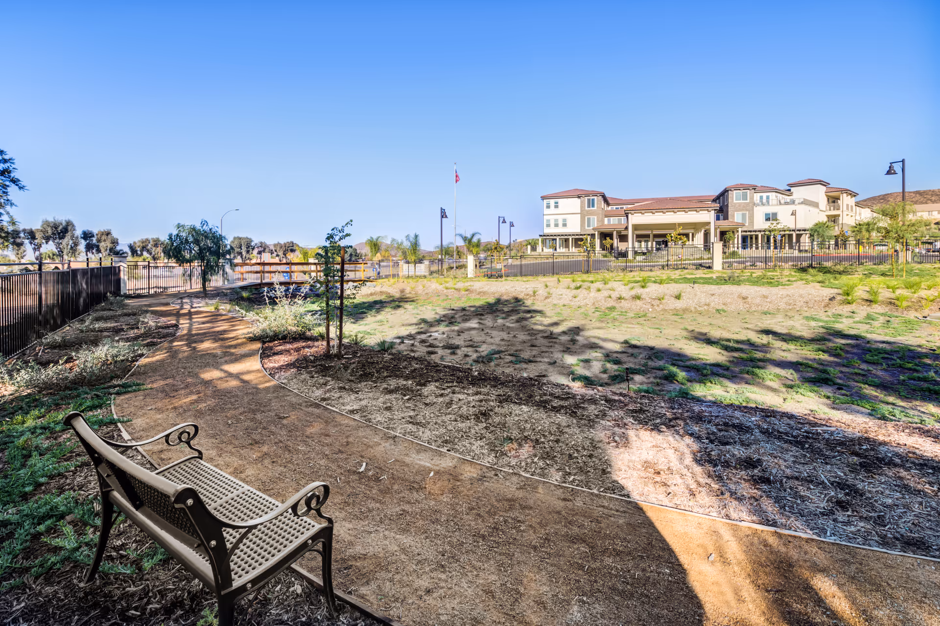 Outdoor walking path with a bench along the side, surrounded by landscaped plants and trees, leading towards a large senior living facility building under a clear blue sky.