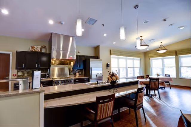 A bright and spacious kitchen and dining area with large windows allowing natural light to fill the room. The kitchen features dark cabinetry, a stainless steel refrigerator, a large stove with a stainless steel hood, and a long countertop with a sink and several chairs. The dining area has tables covered with tablecloths and wooden chairs. The room has wooden flooring and modern ceiling lights.