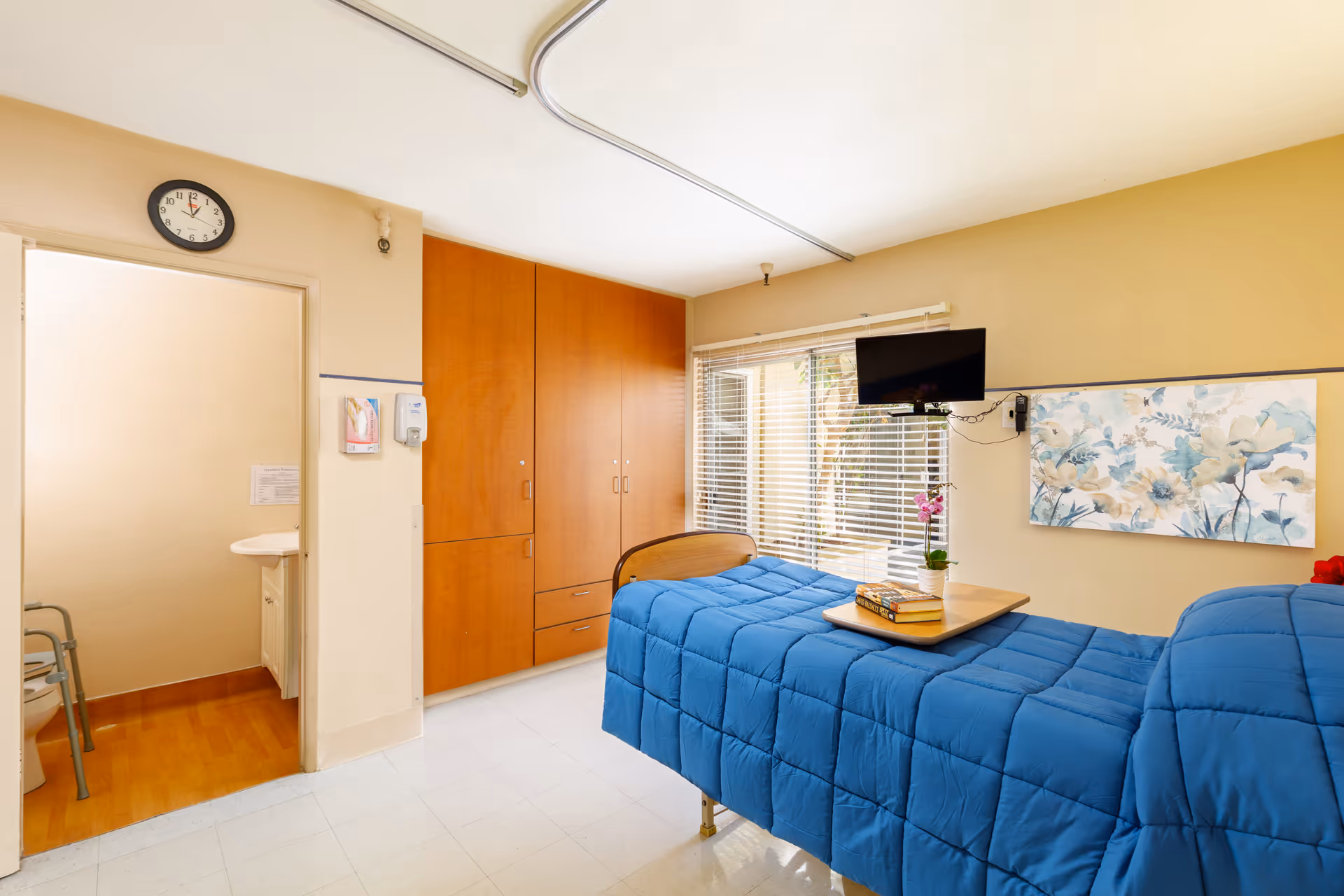 A bright and clean senior living bedroom with a single bed covered in a blue comforter. The room features a wooden wardrobe, a wall-mounted TV, a window with blinds, and a floral painting on the wall. There is a small table on the bed with books and a potted plant. An open door reveals a bathroom with a toilet and a sink.