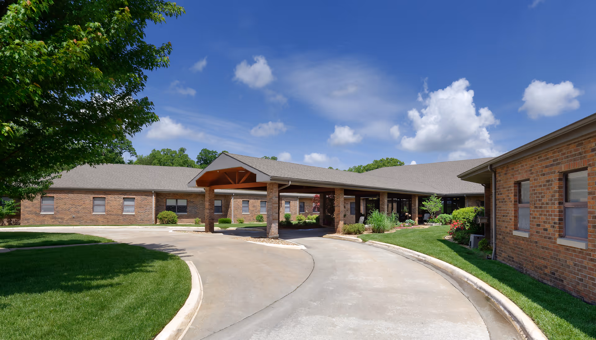 Brick single-story senior care facility with a covered entrance, circular driveway, and landscaped lawn under a blue sky.