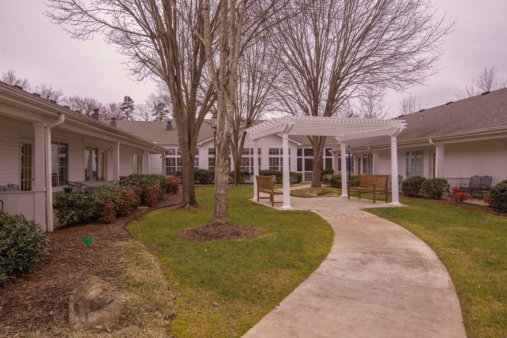 Outdoor courtyard area at The Lantern at Morning Pointe Alzheimer's Center of Excellence Chattanooga featuring a curved concrete pathway, green grass, leafless trees, a white pergola with wooden benches underneath, and surrounding single-story buildings with windows and doors.