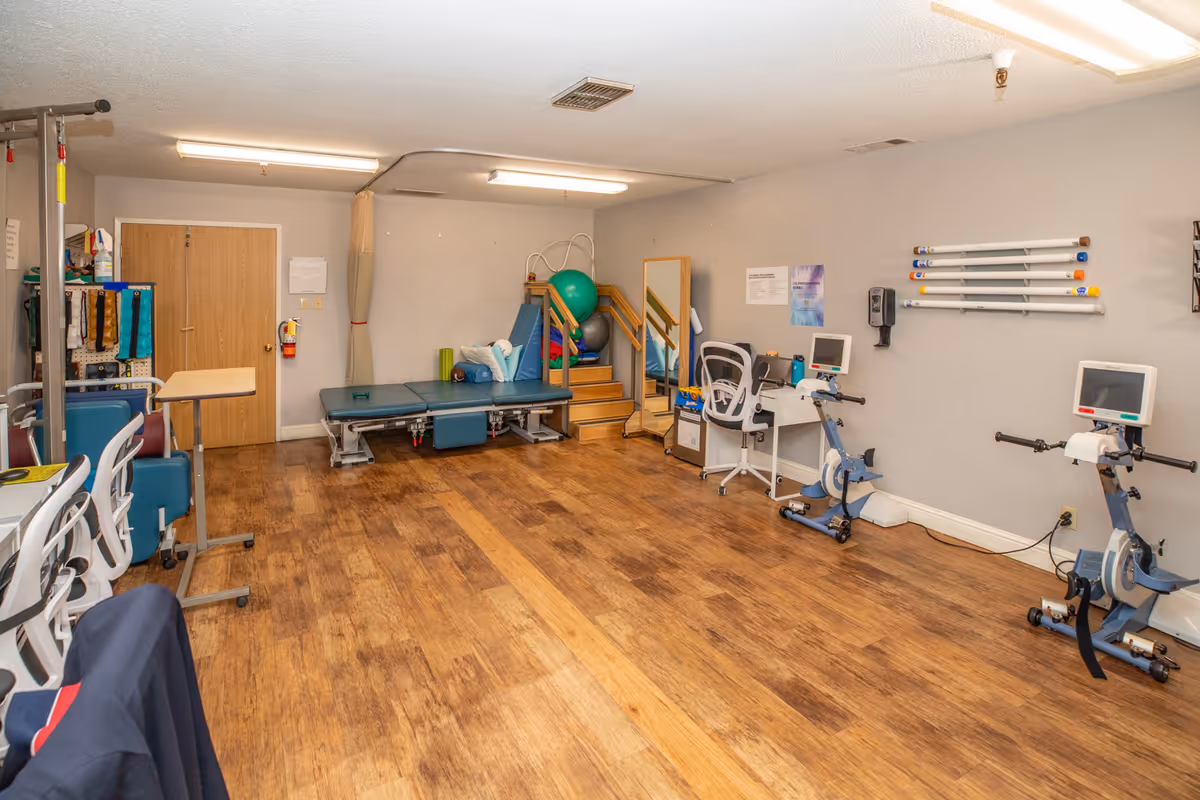 A physical therapy room with wooden flooring, two stationary exercise bikes with monitors, a padded treatment table, therapy balls, a mirror, and various exercise equipment and supplies along the walls.
