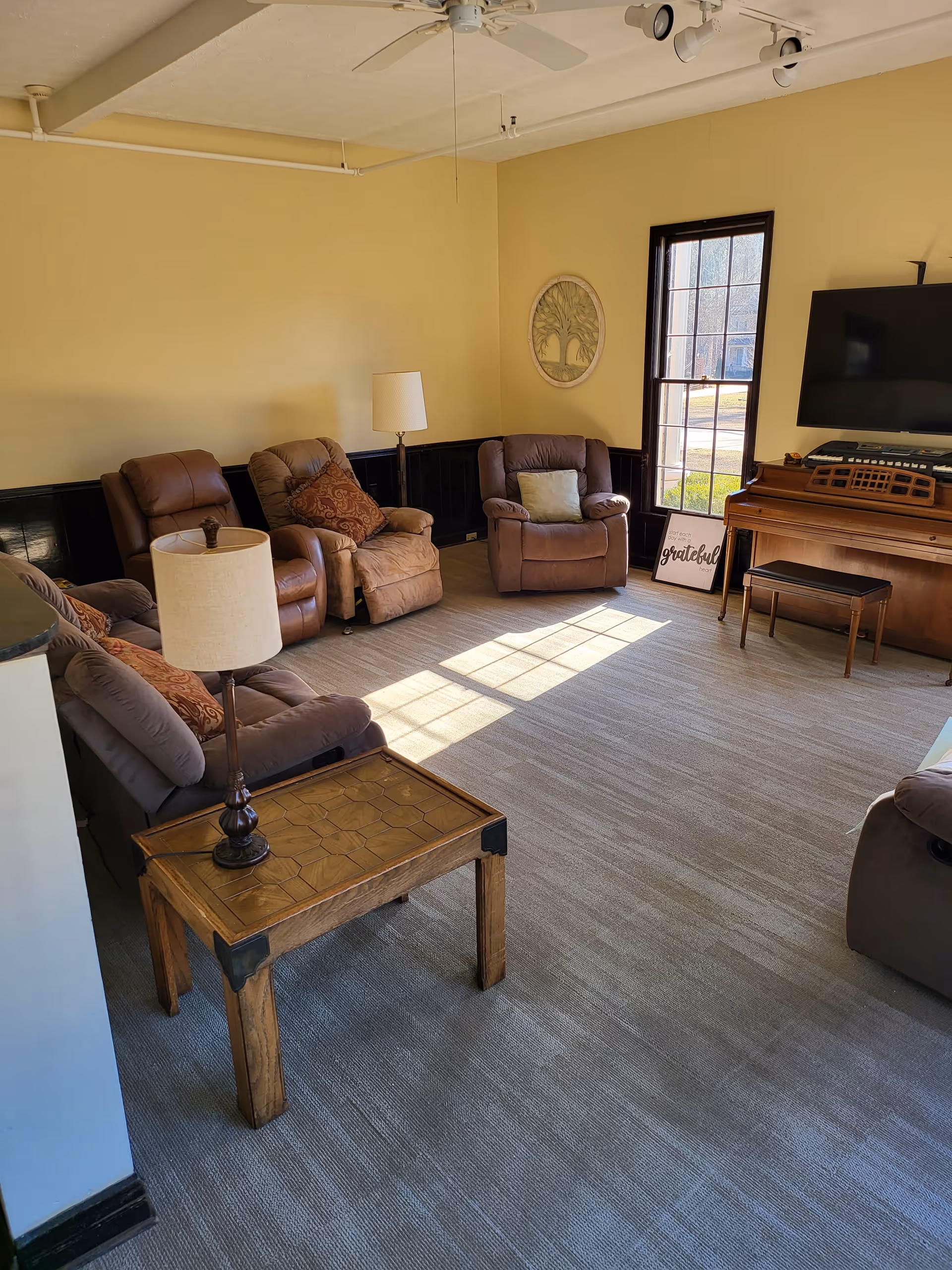 A cozy living room with beige walls and carpeted floor, featuring several cushioned recliner chairs arranged around a wooden side table with a lamp. A large window lets in natural light, illuminating the room. On one wall, there is a flat-screen TV mounted above a wooden piano with a small bench. A decorative wall hanging and a framed sign that reads 'grateful' are also visible.