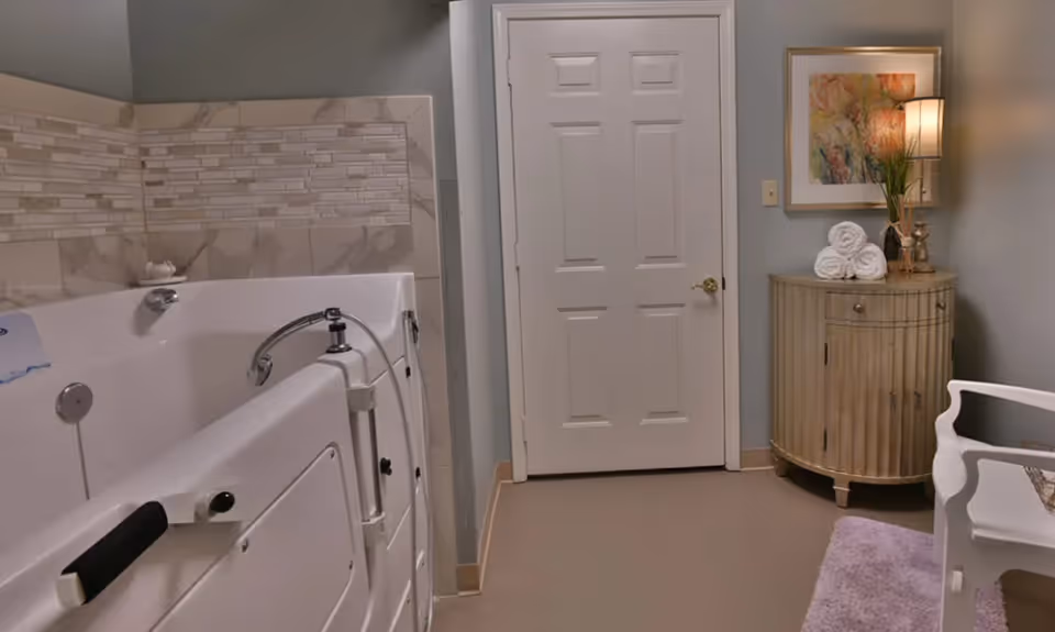 A bathroom with a walk-in bathtub on the left side, a closed white door in the center, and a small wooden cabinet with rolled white towels, a lamp, and a framed artwork on the right. A white chair and a purple rug are partially visible on the right side.