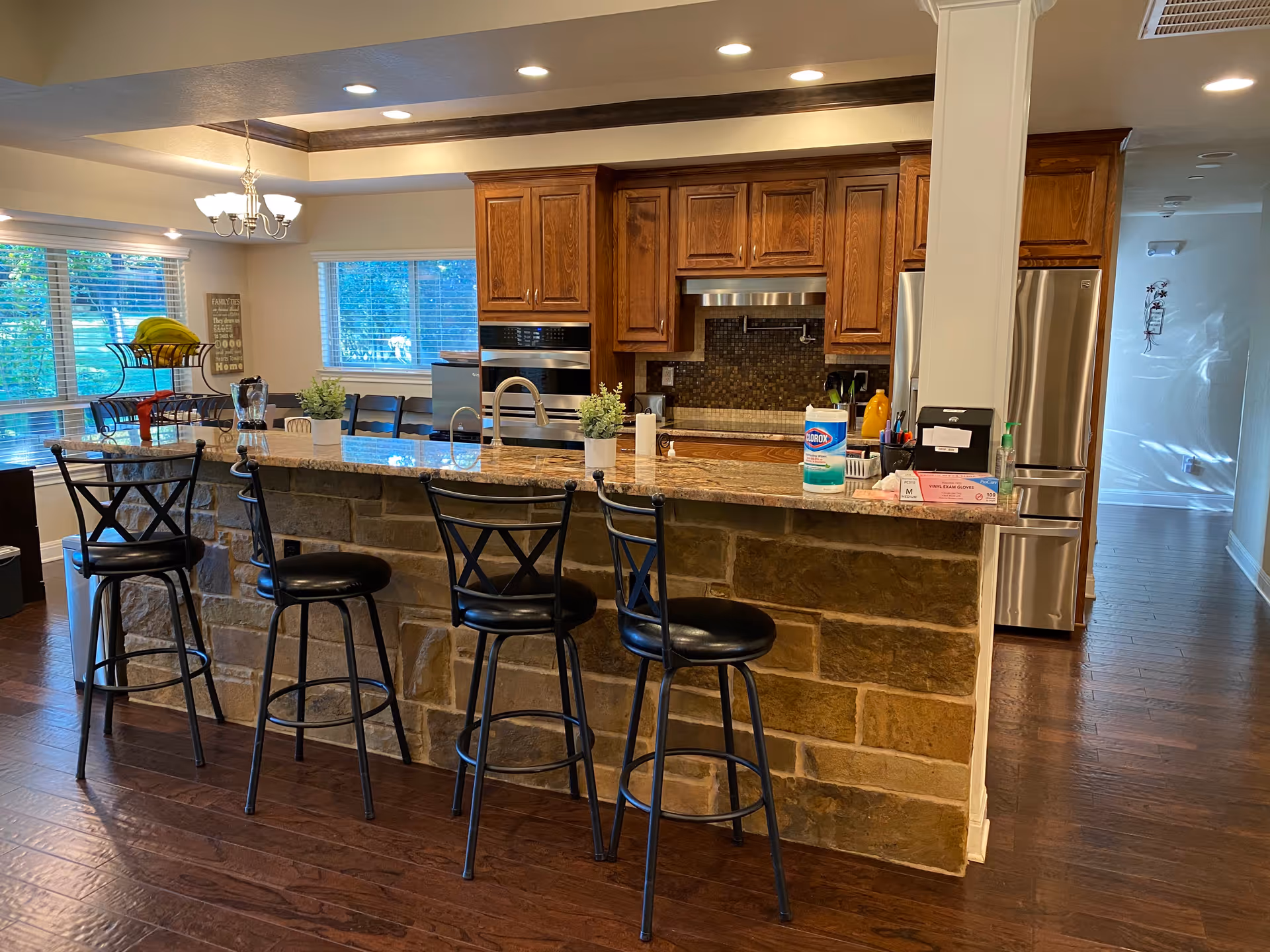 Interior view of a kitchen with a stone and granite island counter featuring four black bar stools. The kitchen has wooden cabinets, stainless steel appliances including an oven and refrigerator, and a tiled backsplash. There are windows with blinds letting in natural light, a chandelier light fixture, and various items on the counter such as cleaning wipes, a blender, and a fruit basket.