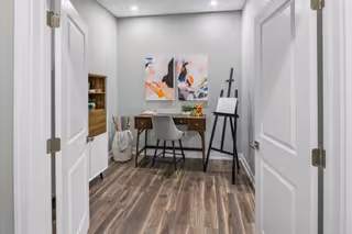 Small interior art studio/office with a desk, chair, easel, decorative wall art and wood-look flooring seen through open double doors.