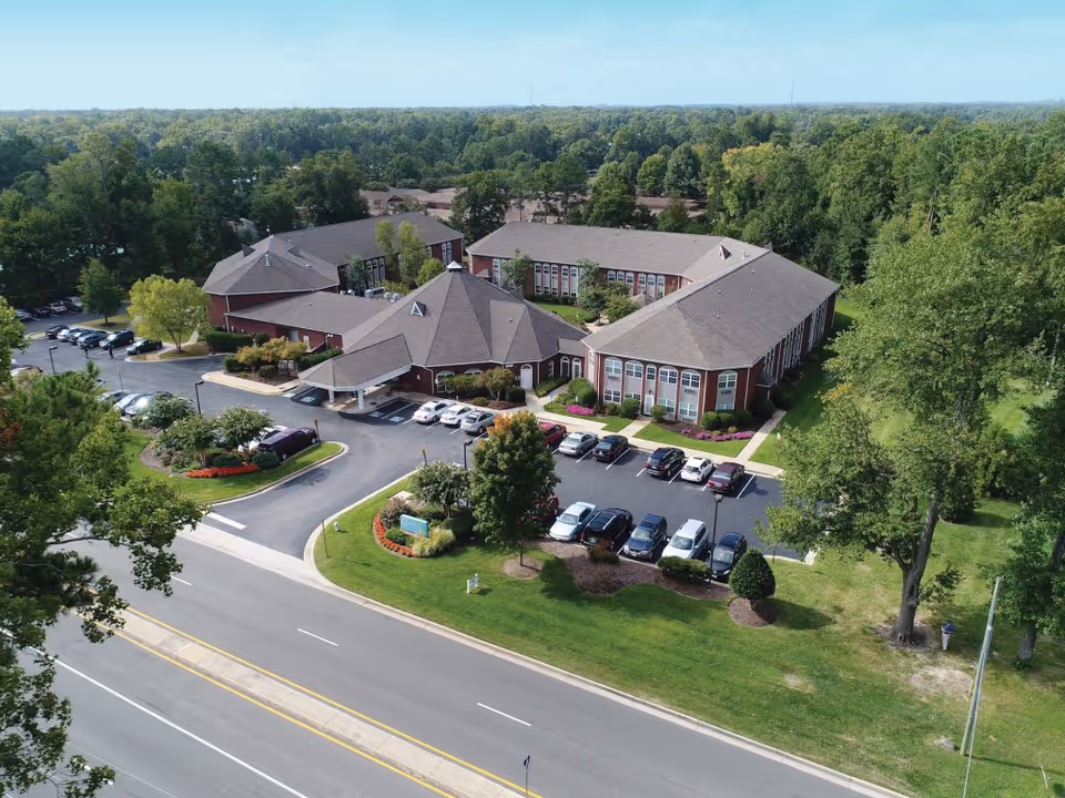 Aerial view of Chestnut Grove Assisted Living facility showing a large, single-story building with multiple wings, surrounded by trees and greenery. There is a parking lot with several cars parked, landscaped areas with bushes and flowers, and a road in the foreground.