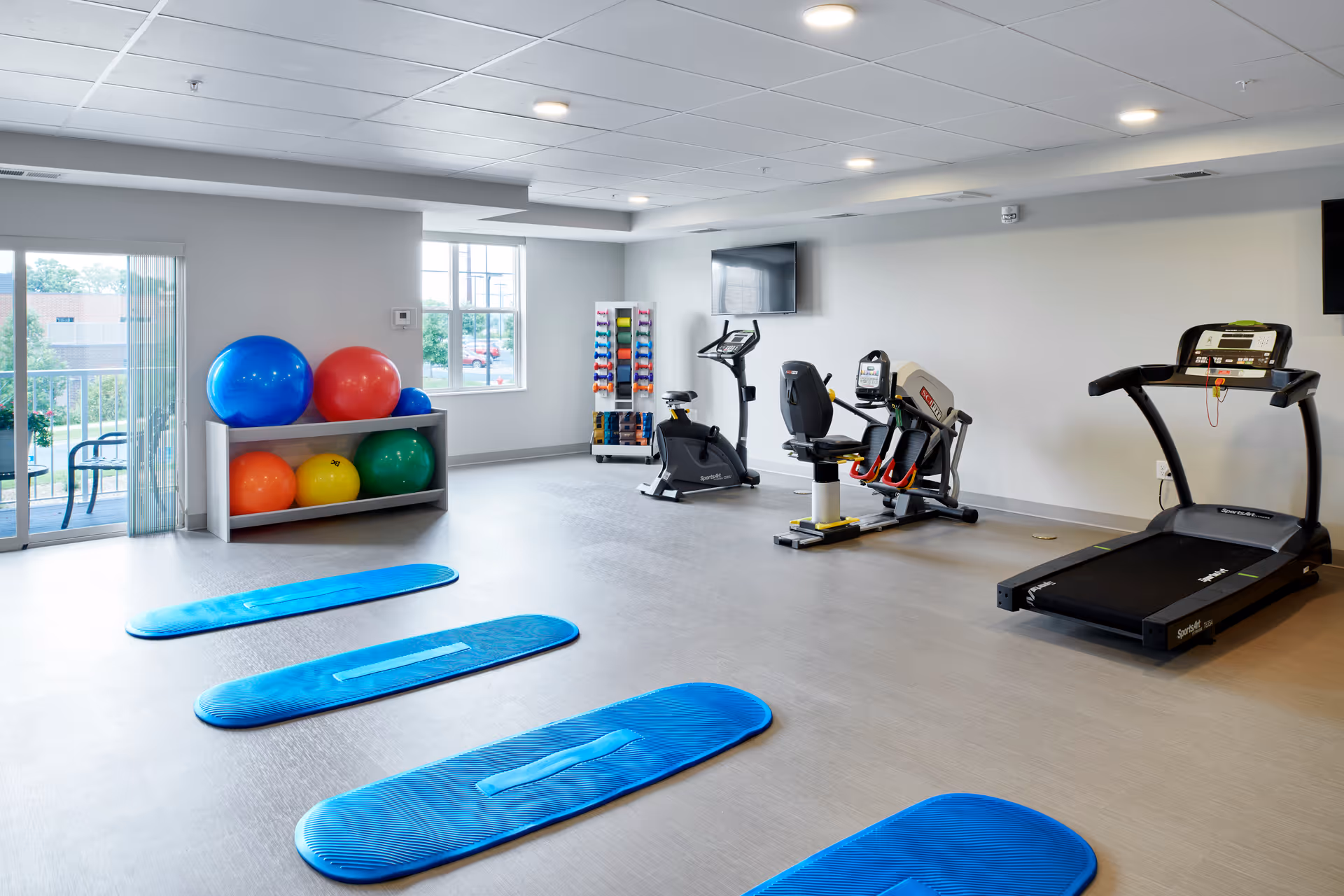 A bright fitness room with exercise equipment including a treadmill, stationary bike, and leg press machine. There are three blue exercise mats laid out on the floor, a rack with colorful dumbbells, and a shelf holding large exercise balls in various colors. Large windows and a glass door let in natural light.