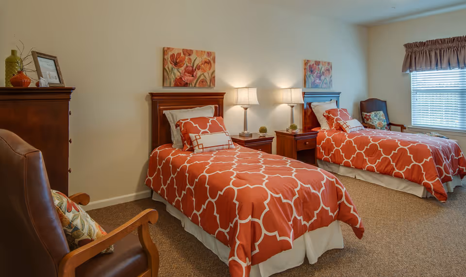A senior living bedroom with two twin beds featuring orange and white patterned bedspreads. Each bed has pillows and a wooden headboard. Between the beds is a wooden nightstand with two lamps and decorative items. There is a wooden dresser on the left side and a brown armchair with a floral cushion. A window with blinds and a valance is on the right wall, and floral paintings hang above each bed.