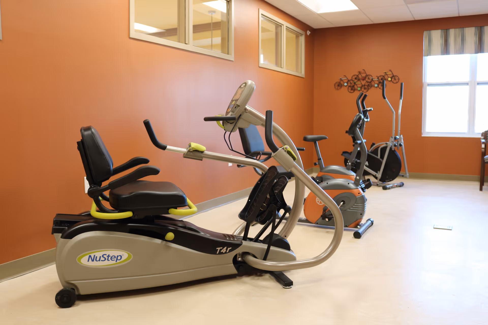 A small fitness room with several exercise bikes against an orange wall and a window.