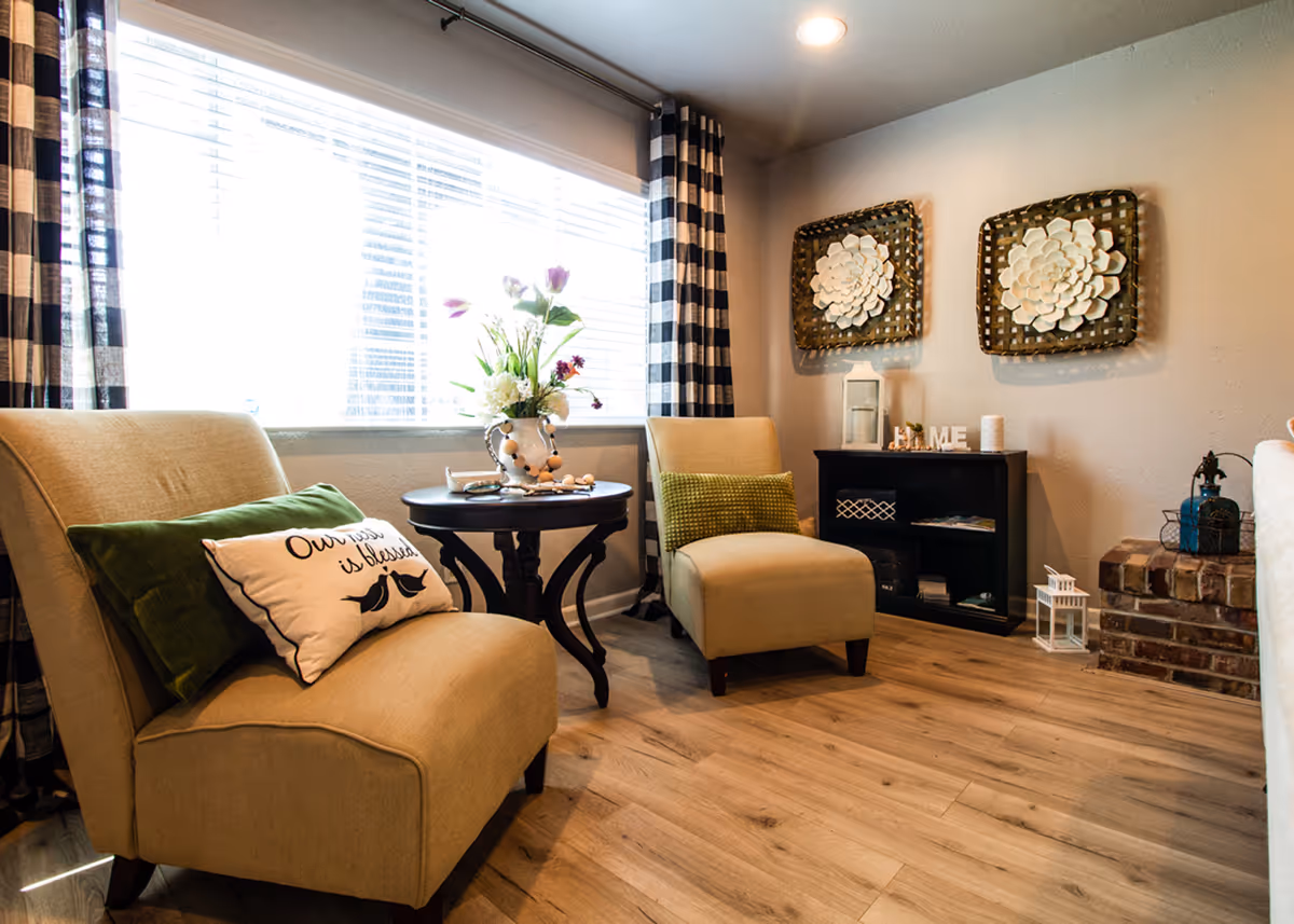 Bright, cozy living room with two beige chairs, a round side table holding flowers, plaid curtains, decorative wall art, and a small shelving unit.