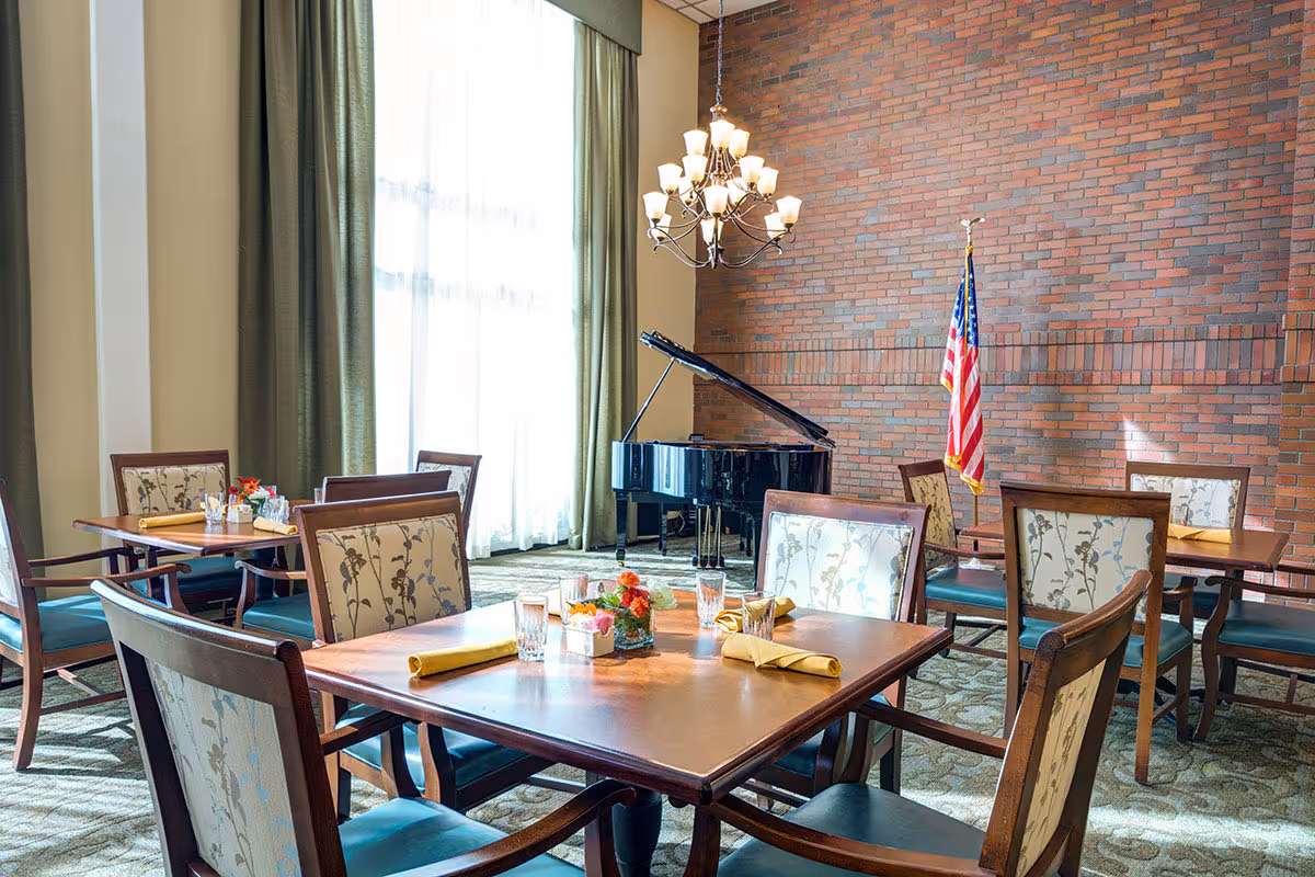 Bright dining room with tables set for meals, a grand piano, chandelier, and an American flag against a brick wall.