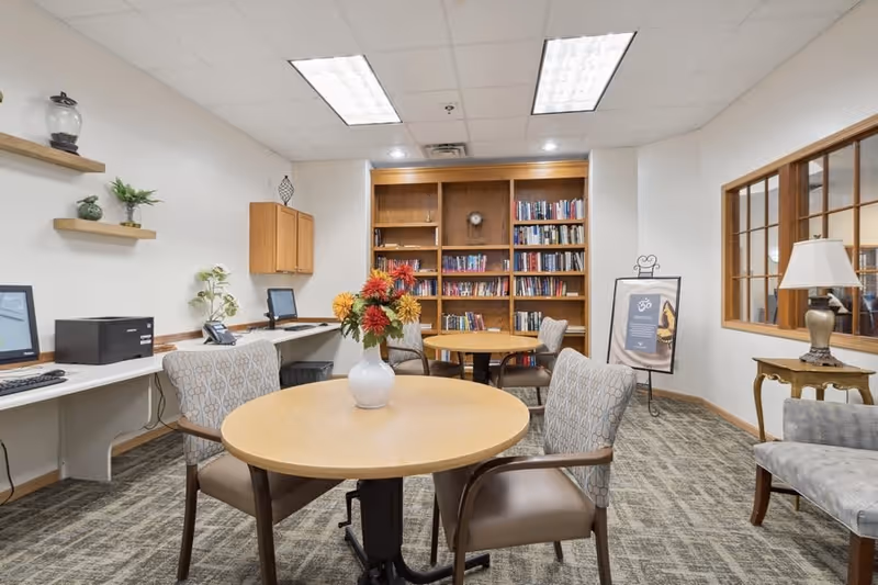 A cozy interior room with round wooden tables and cushioned chairs arranged for seating. A vase with colorful flowers is placed on the nearest table. In the background, there is a wooden bookshelf filled with books, a framed poster on an easel, and a window with wooden trim looking into another room. On the left side, there are computer workstations with monitors, a printer, and decorative shelves with plants and ornaments on the wall. The room has a carpeted floor and a white ceiling with fluorescent lights.