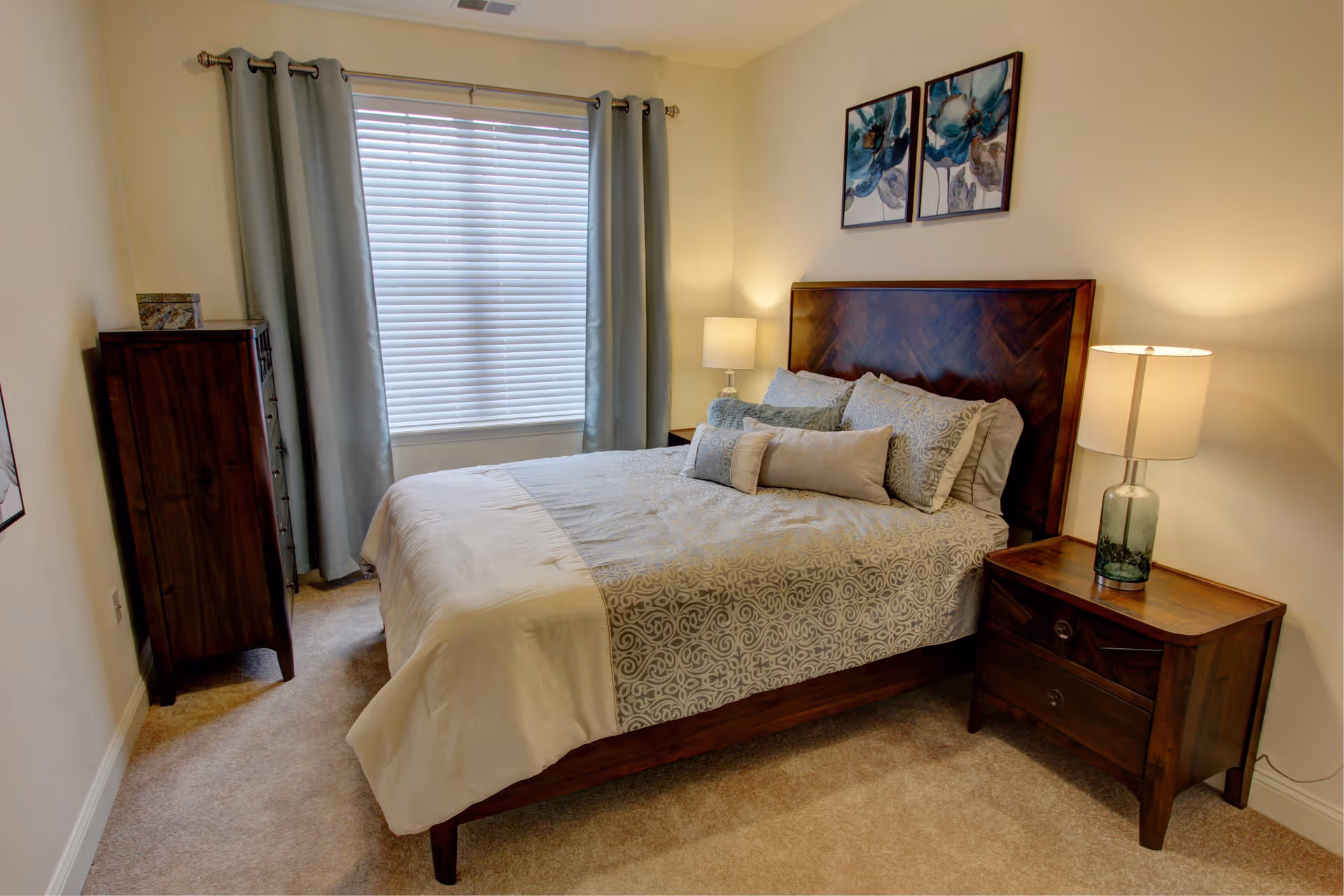 A neatly made bed with patterned bedding and multiple pillows in a bedroom. The room features a window with closed blinds and light blue curtains, a wooden nightstand with a lamp on the right side of the bed, a tall wooden dresser on the left side near the window, and two floral paintings hanging above the headboard.
