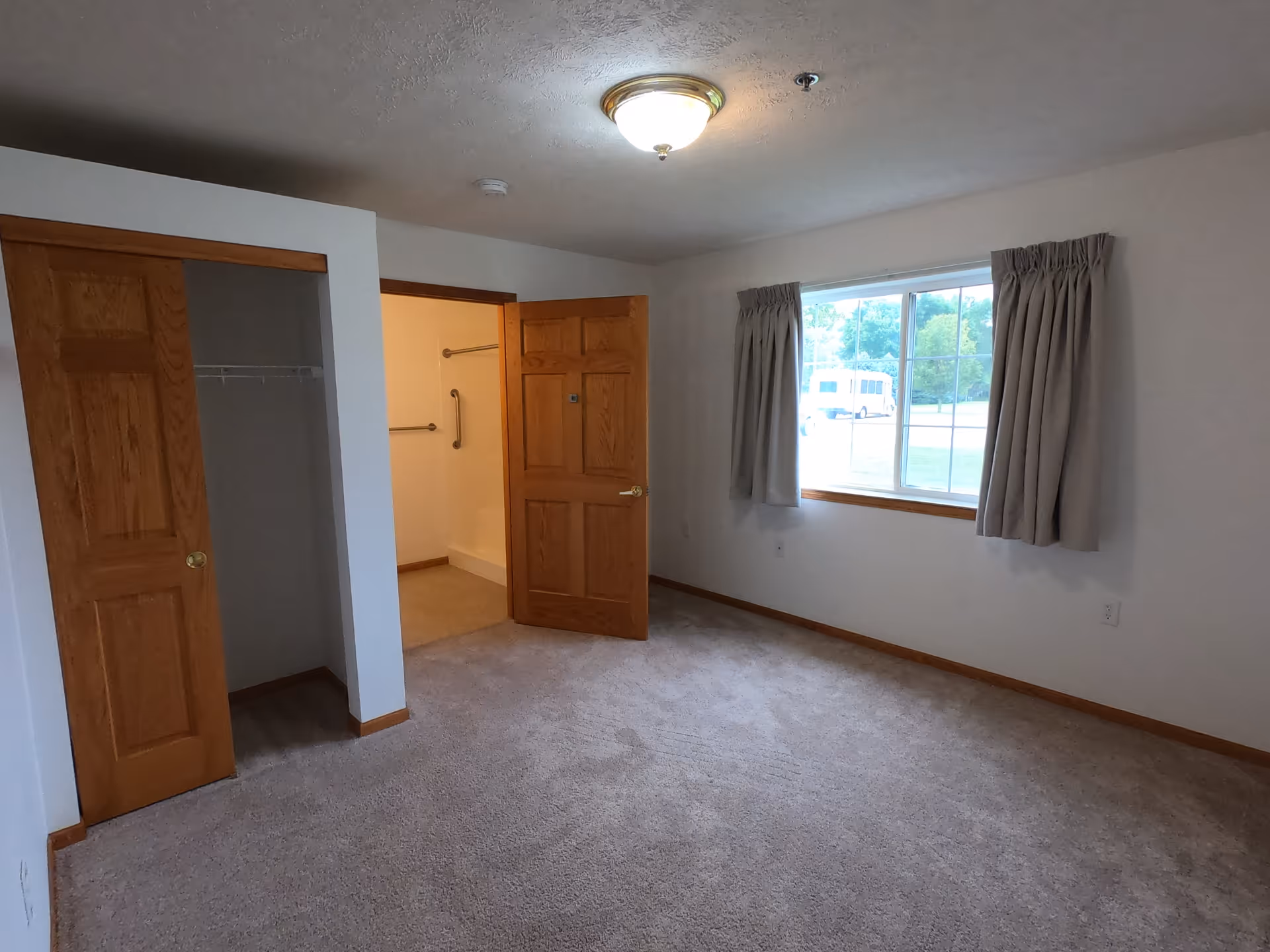 Empty room with beige carpet, white walls, a window with beige curtains, a wooden closet with sliding doors, and an open wooden door leading to a bathroom with grab bars.