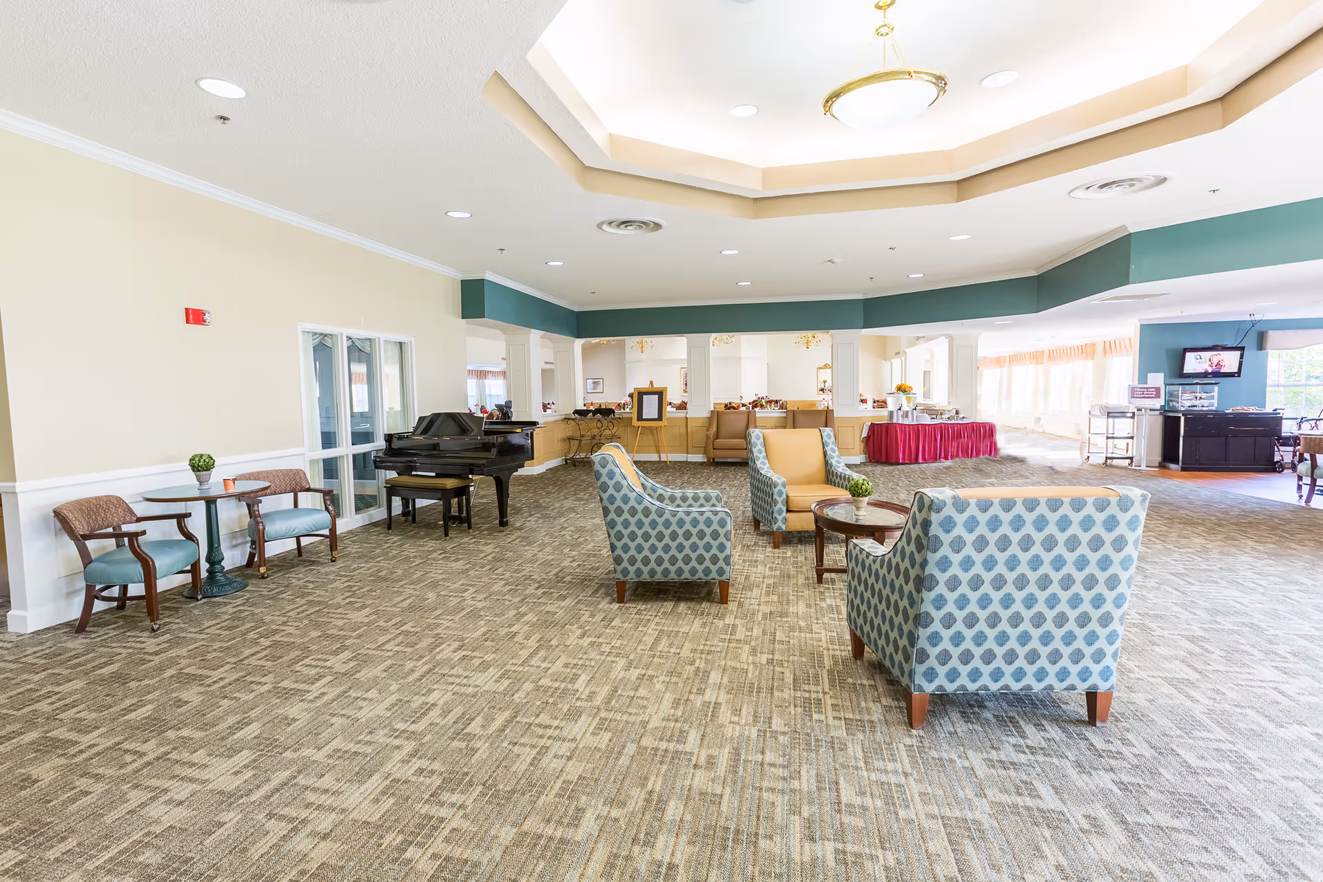 Bright, spacious senior living lounge with upholstered chairs around small tables, a grand piano, and a reception/dining area in the background.