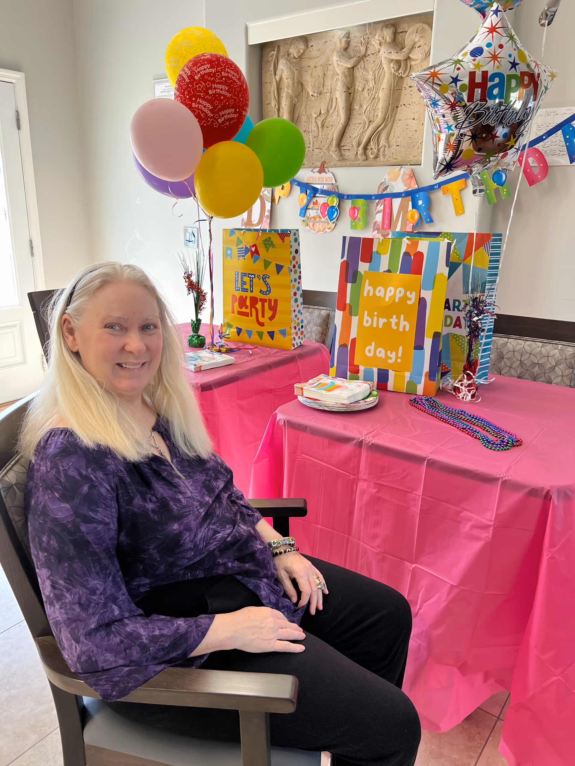 A woman with long blonde hair wearing a purple blouse and black pants is sitting on a chair next to a table covered with a pink tablecloth. The table is decorated with colorful birthday balloons, gift bags, and party napkins. A 'Happy Birthday' banner is hanging on the wall behind the table.