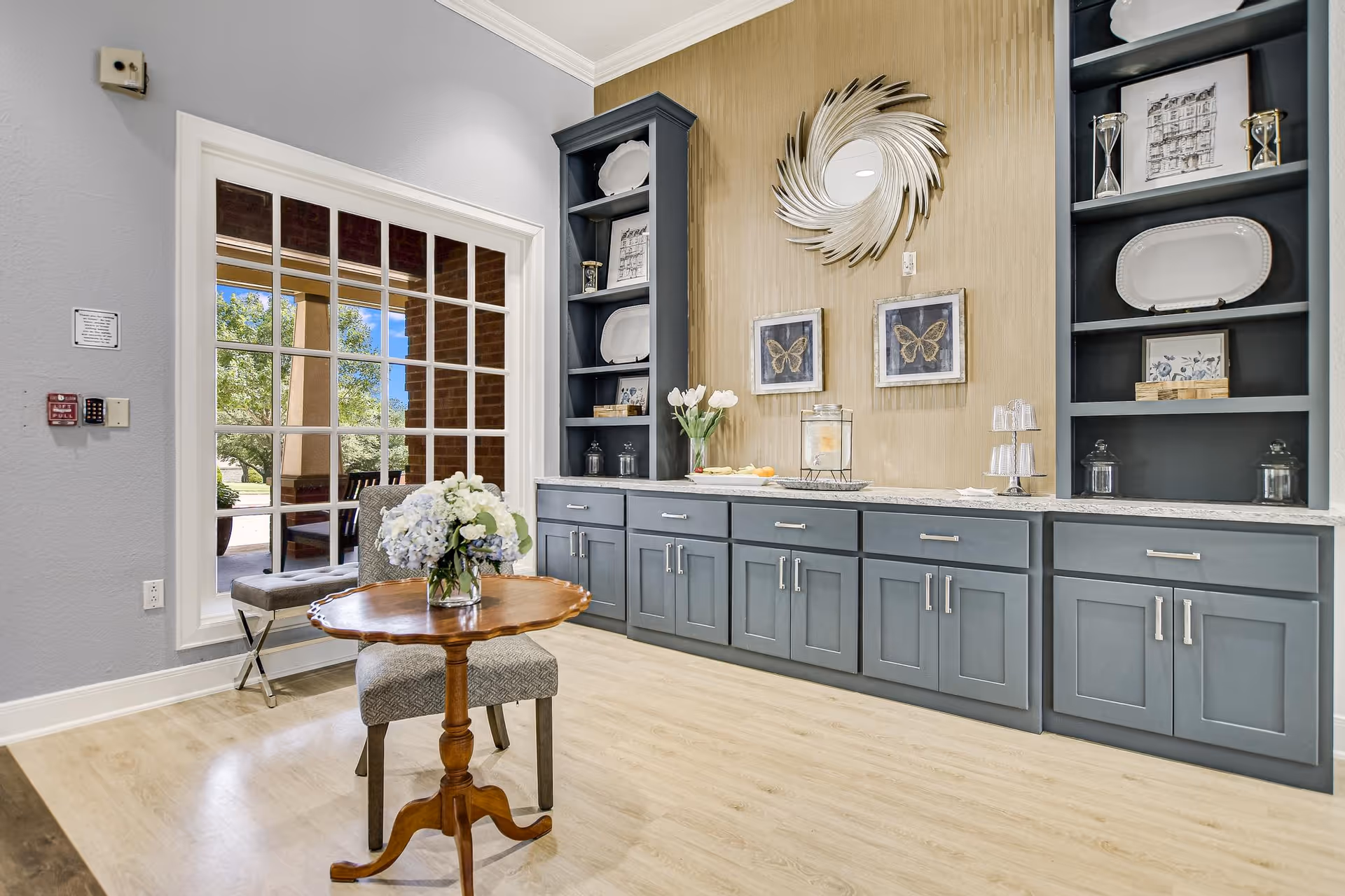 Bright furnished common area featuring a long gray built-in cabinet and shelves, a small round table with a flower arrangement, and a large multi-pane window showing outdoor greenery.