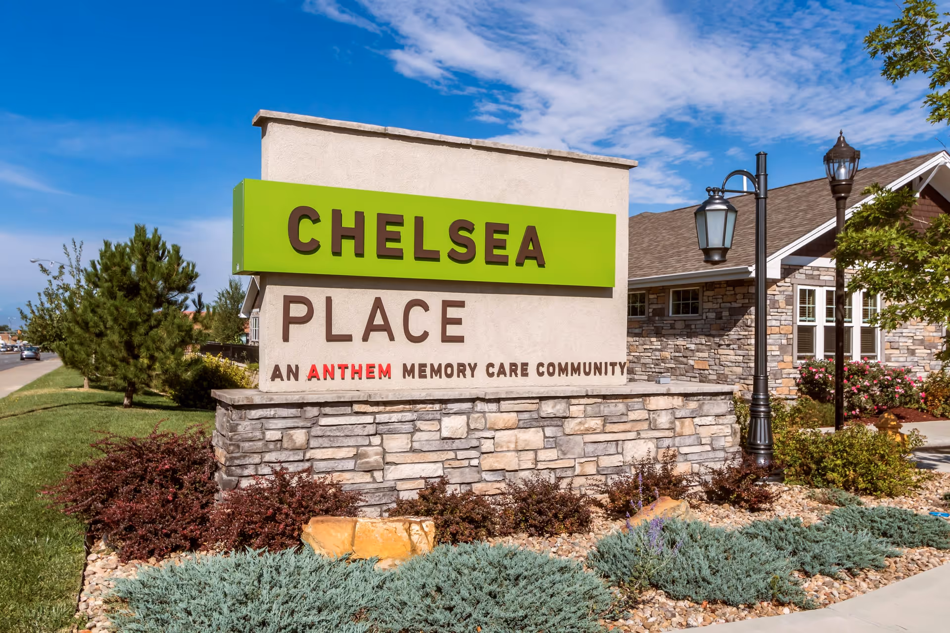 Outdoor view of a stone and stucco sign for Chelsea Place, an Anthem Memory Care Community, with landscaping and a building in the background under a blue sky with some clouds.