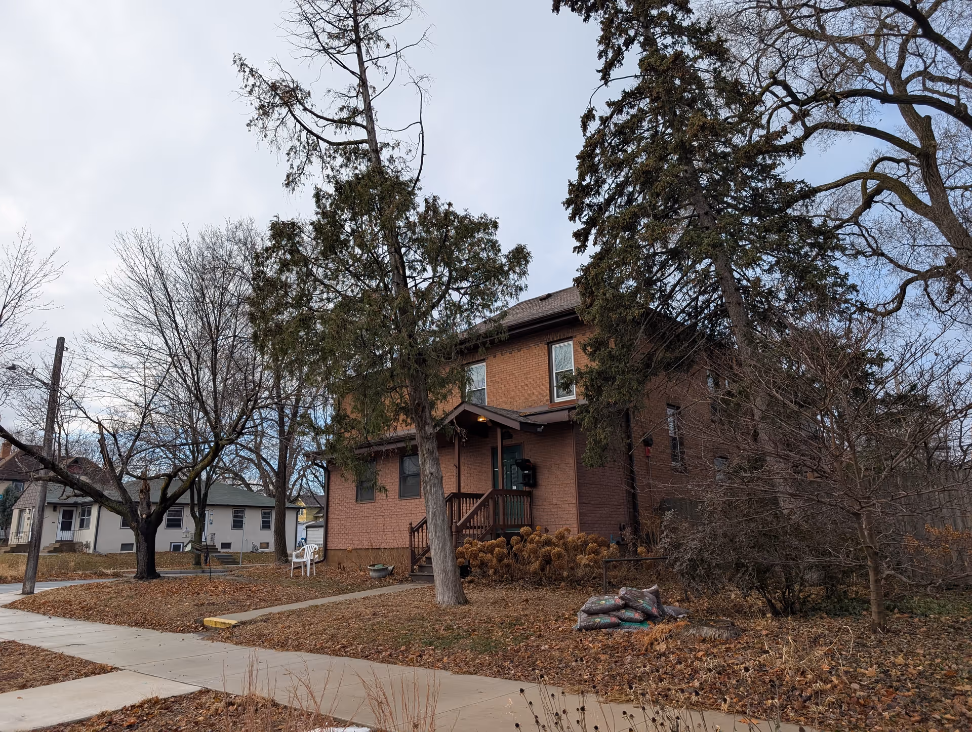 A two-story brick house with a small porch and stairs leading to the front door, surrounded by leafless trees and dry foliage in a residential neighborhood under a cloudy sky.
