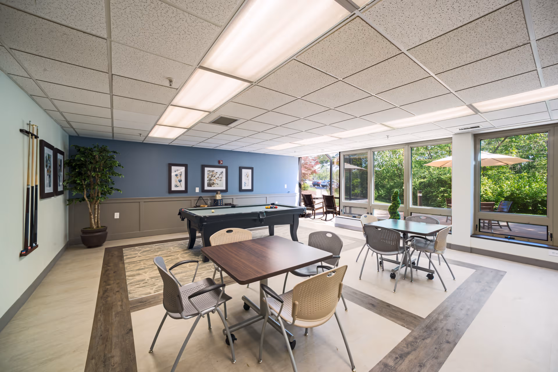 Bright senior community common room with tables and chairs, a pool table, and large windows overlooking a patio.